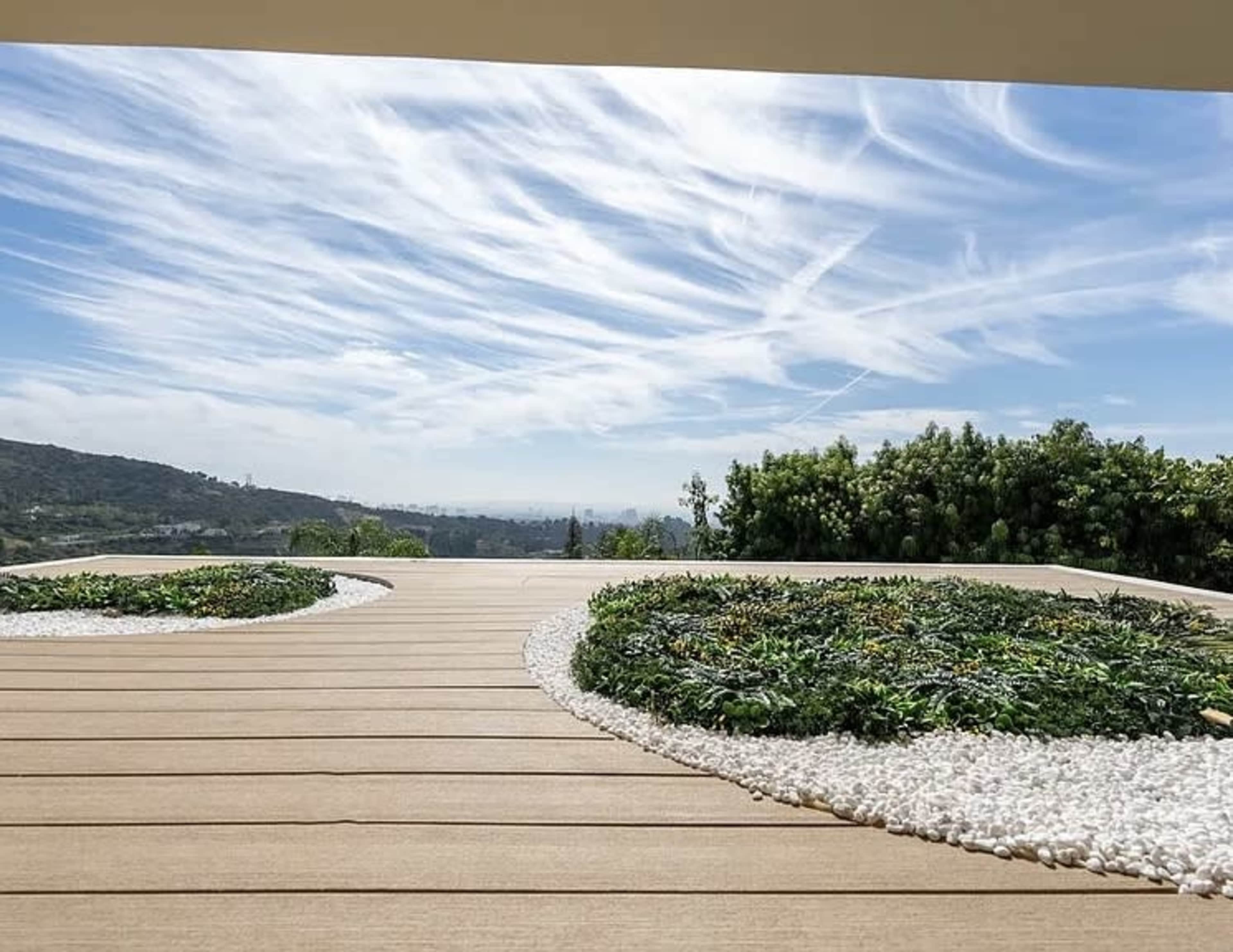 The image shows a wooden deck with circular patches of greenery and white stones, set against a backdrop of cloudy skies and a hilly landscape.