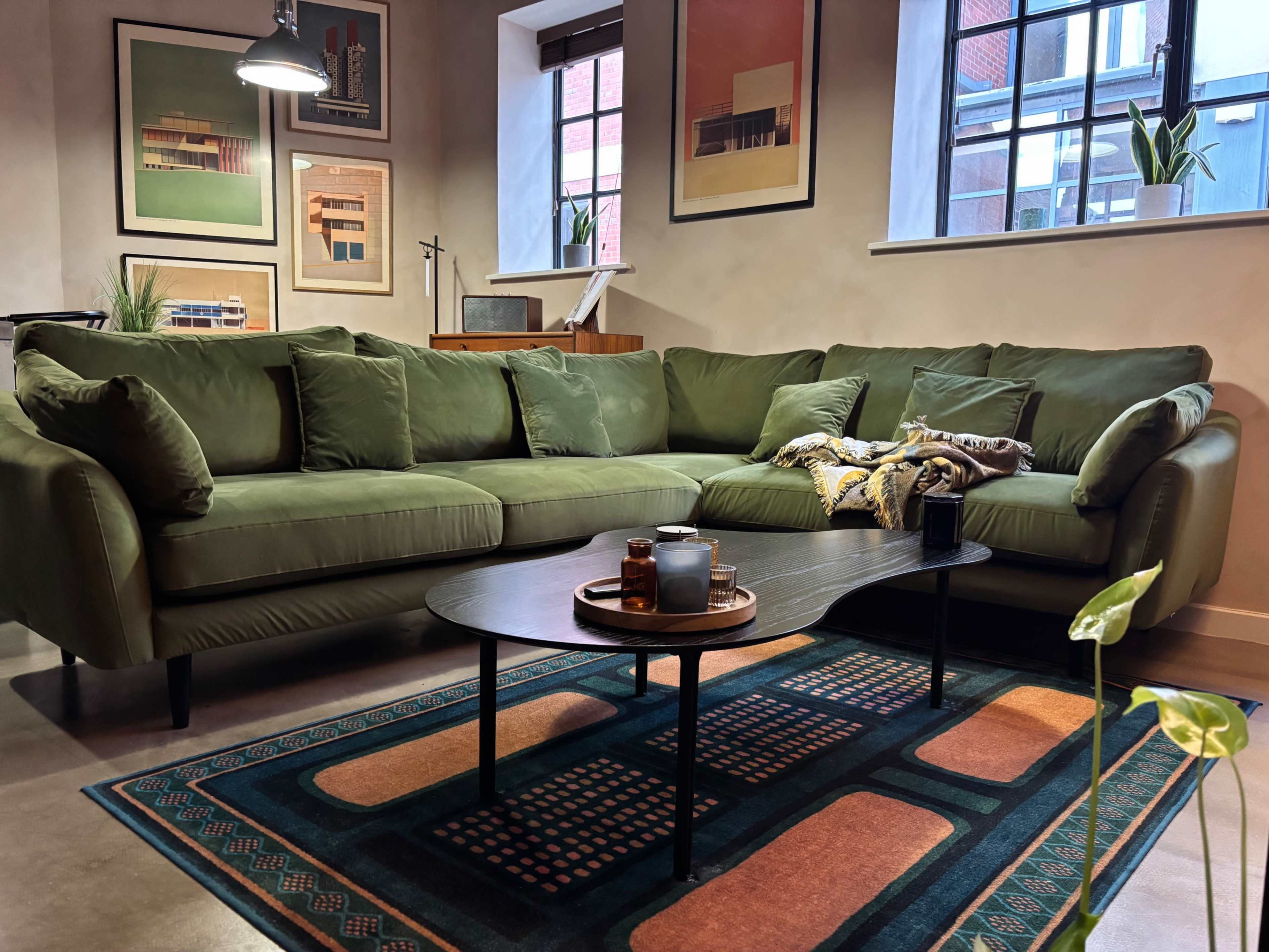 The image features a modern living room with a green sectional sofa, a round coffee table on a patterned rug, and framed art on the walls.