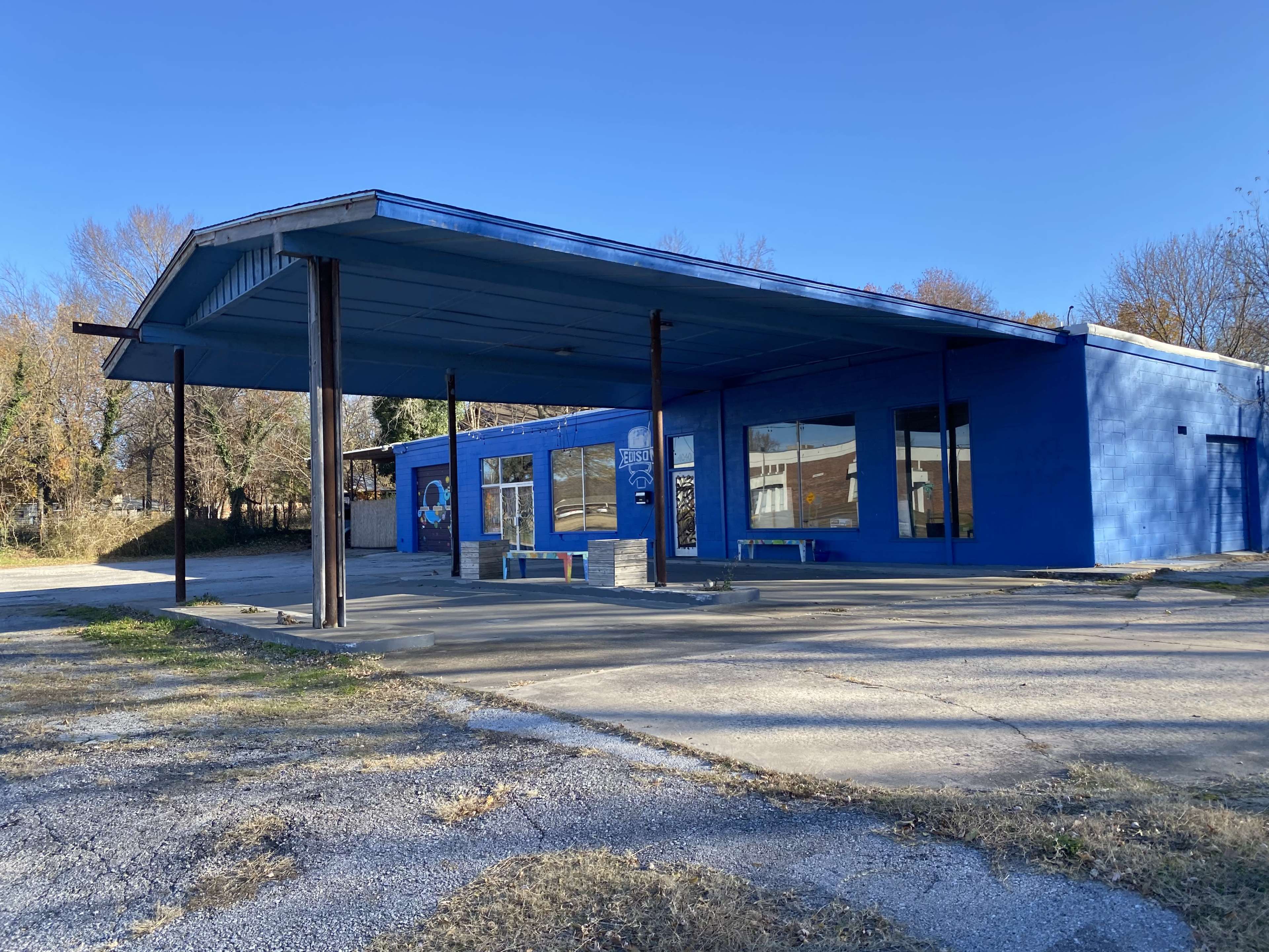A blue-painted building with a canopy and large windows stands on a gravel lot, surrounded by trees.