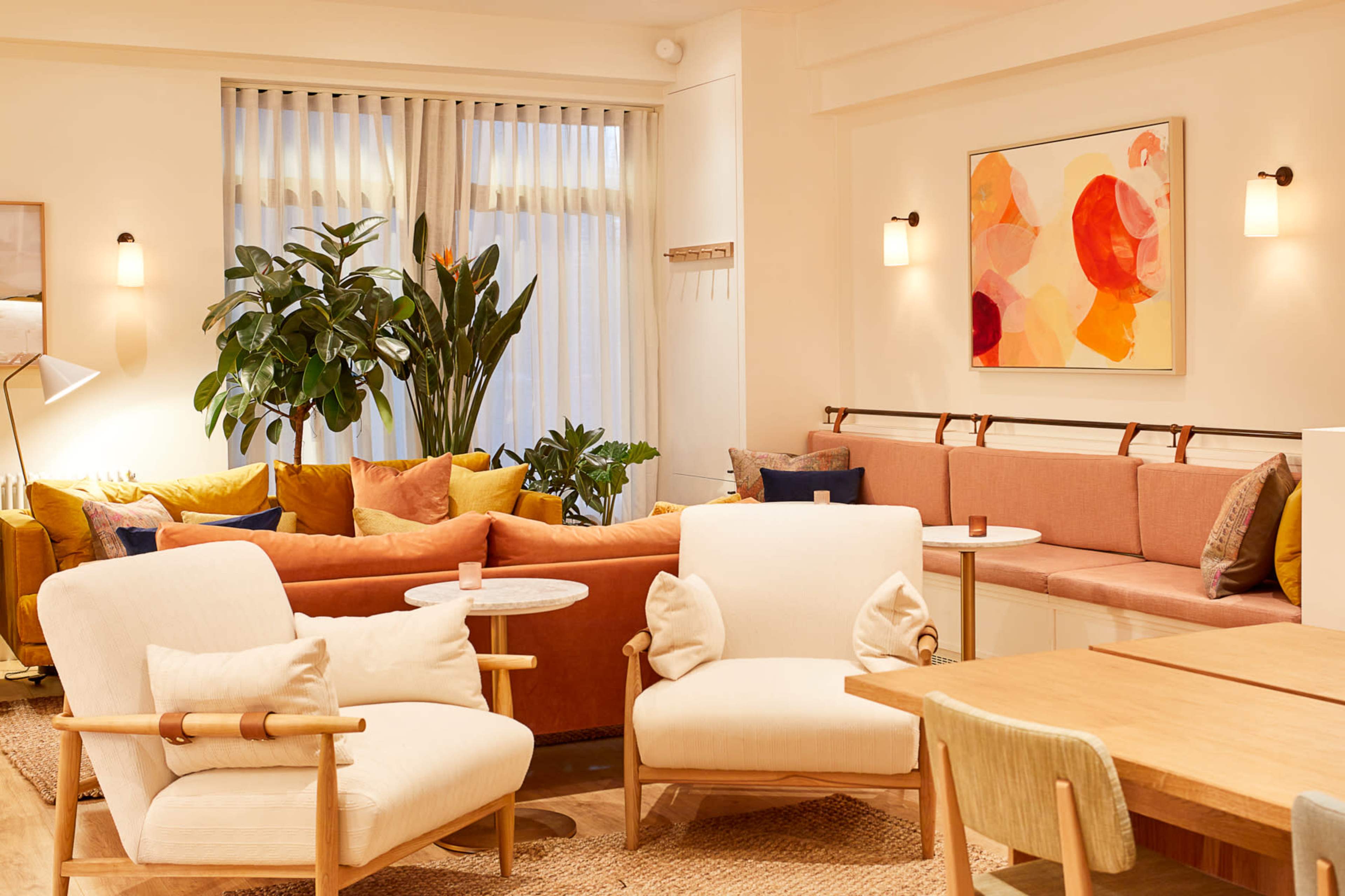 A cozy living space with a mix of seating areas, including armchairs and a couch, surrounded by plants and decorated with colorful cushions and artwork.