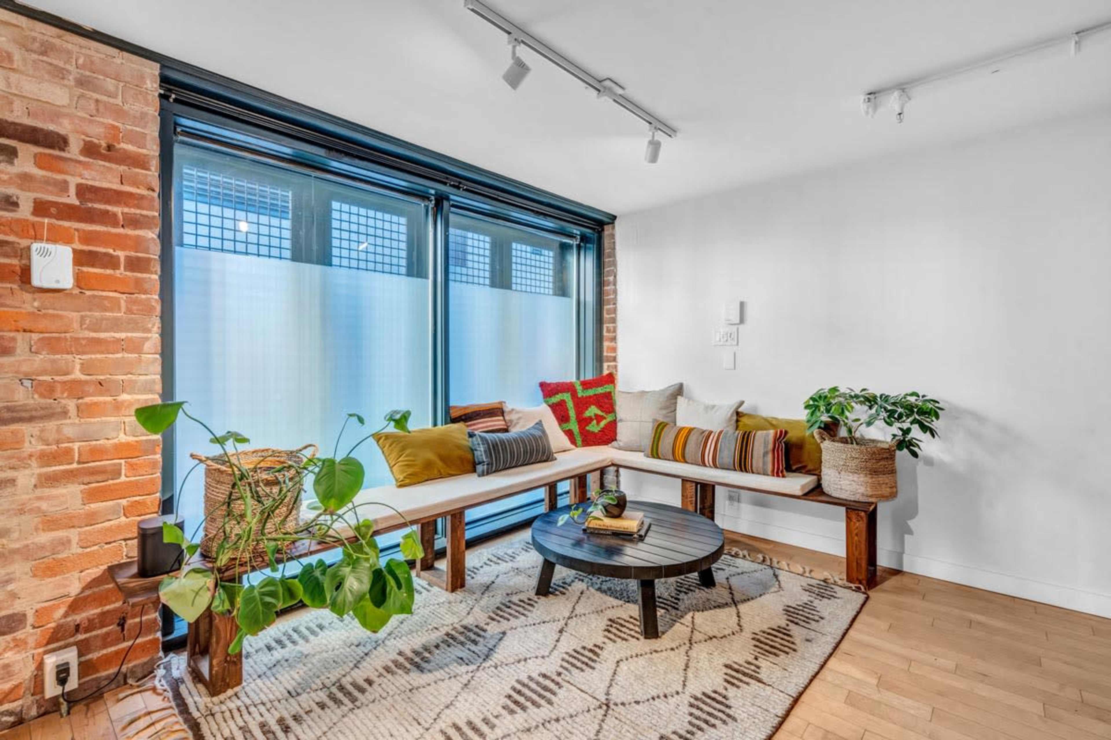 A cozy living space featuring a seating area with a wooden bench, a round black table, and greenery, set against a backdrop of exposed brick walls and large windows.