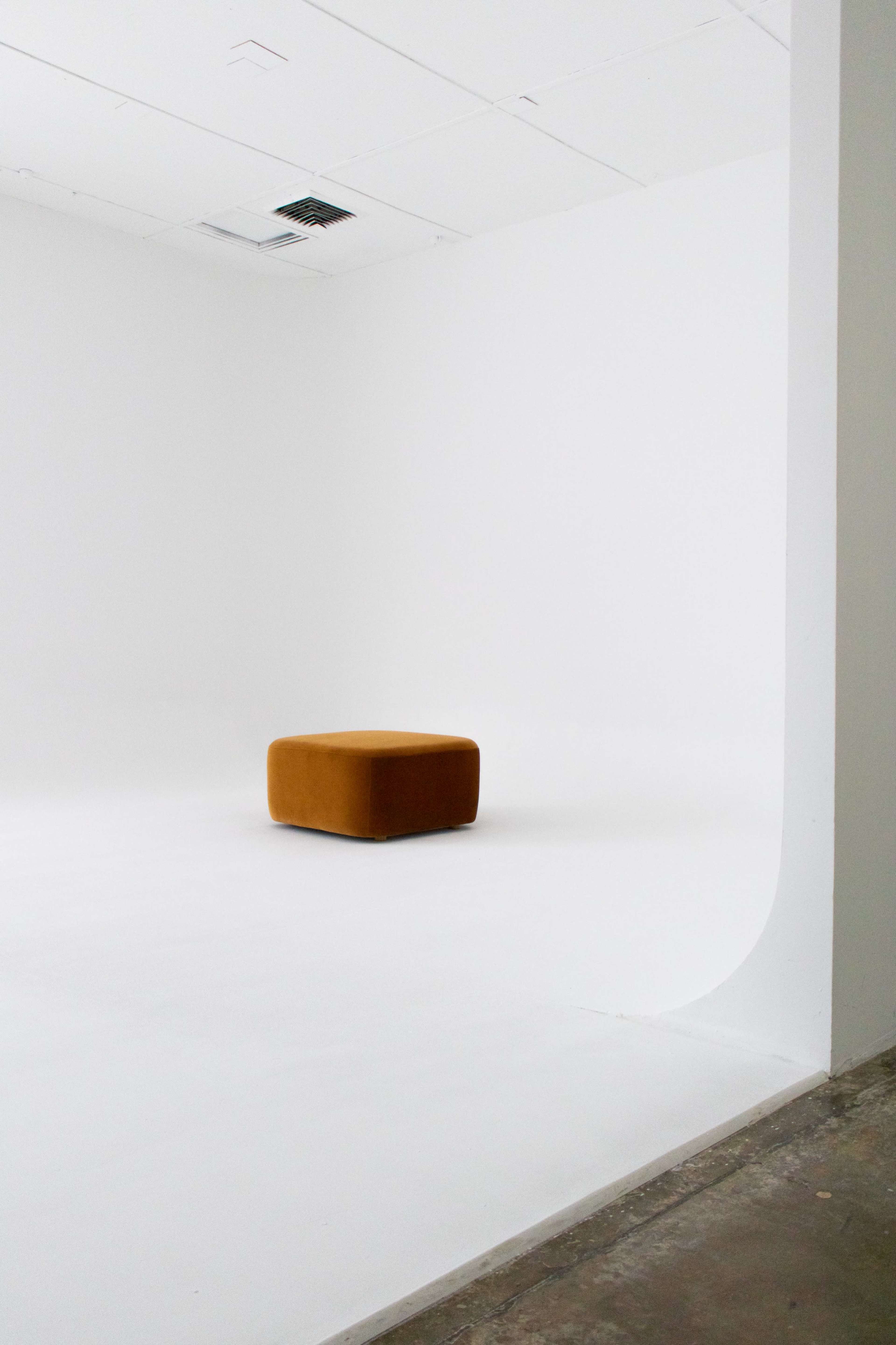 A single brown ottoman is centered in a minimalist, empty white room with smooth walls and a concrete floor.