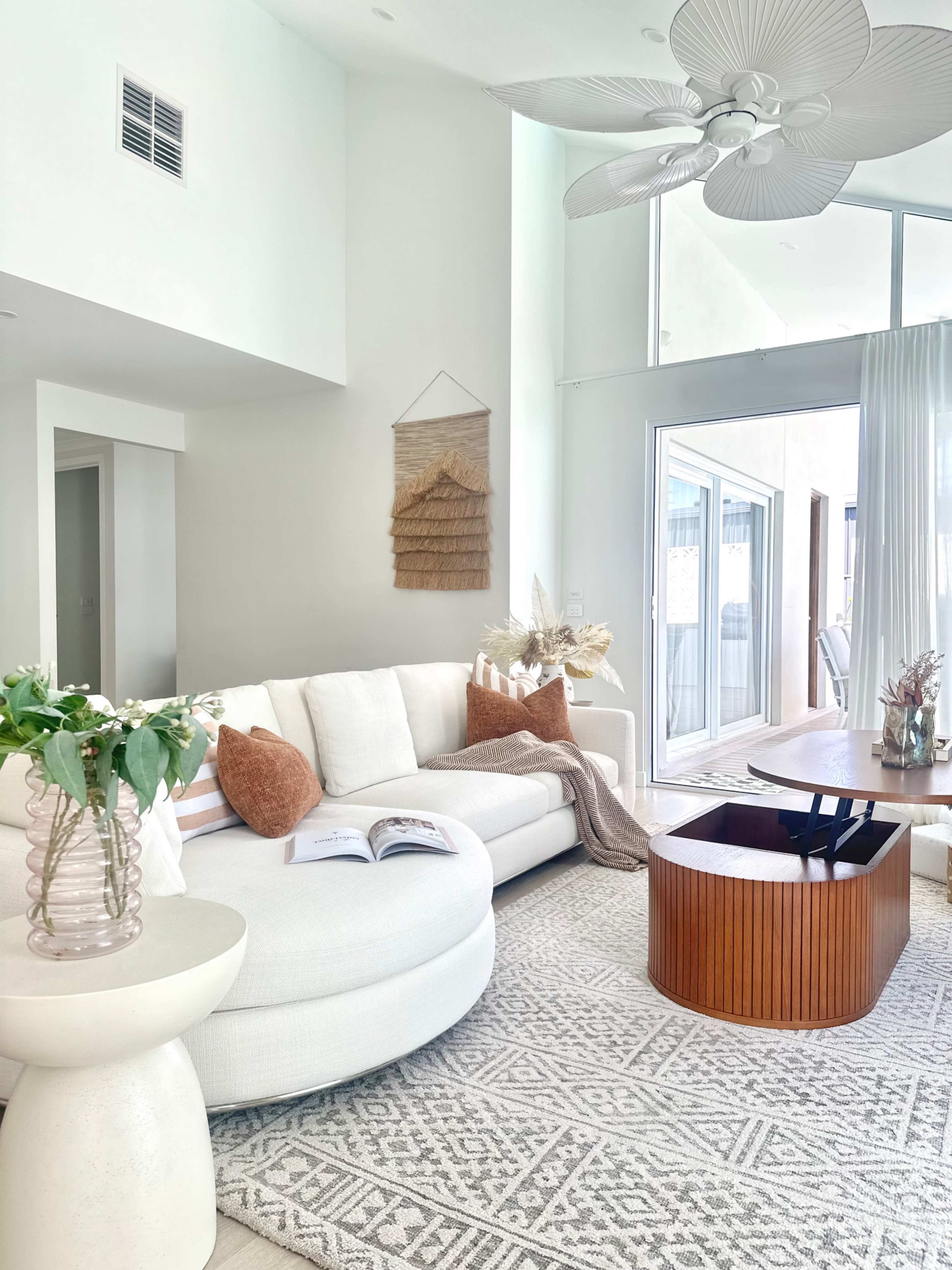 The living room features a white sectional couch, a wooden coffee table, and large windows allowing natural light to enter.