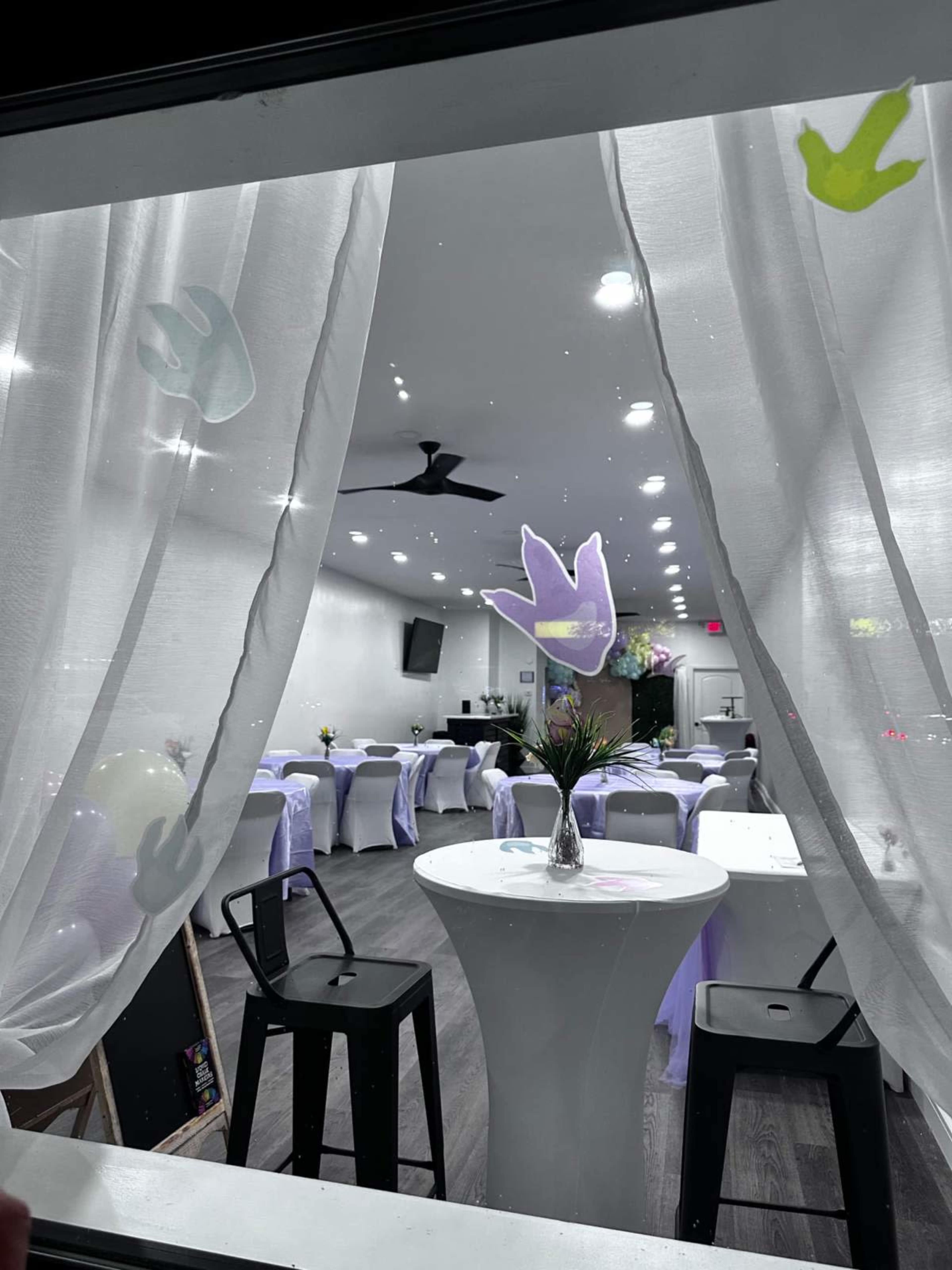 A view through a window of a brightly lit event space with white tables and chairs, featuring light purple decorations and a floral arrangement in the center.