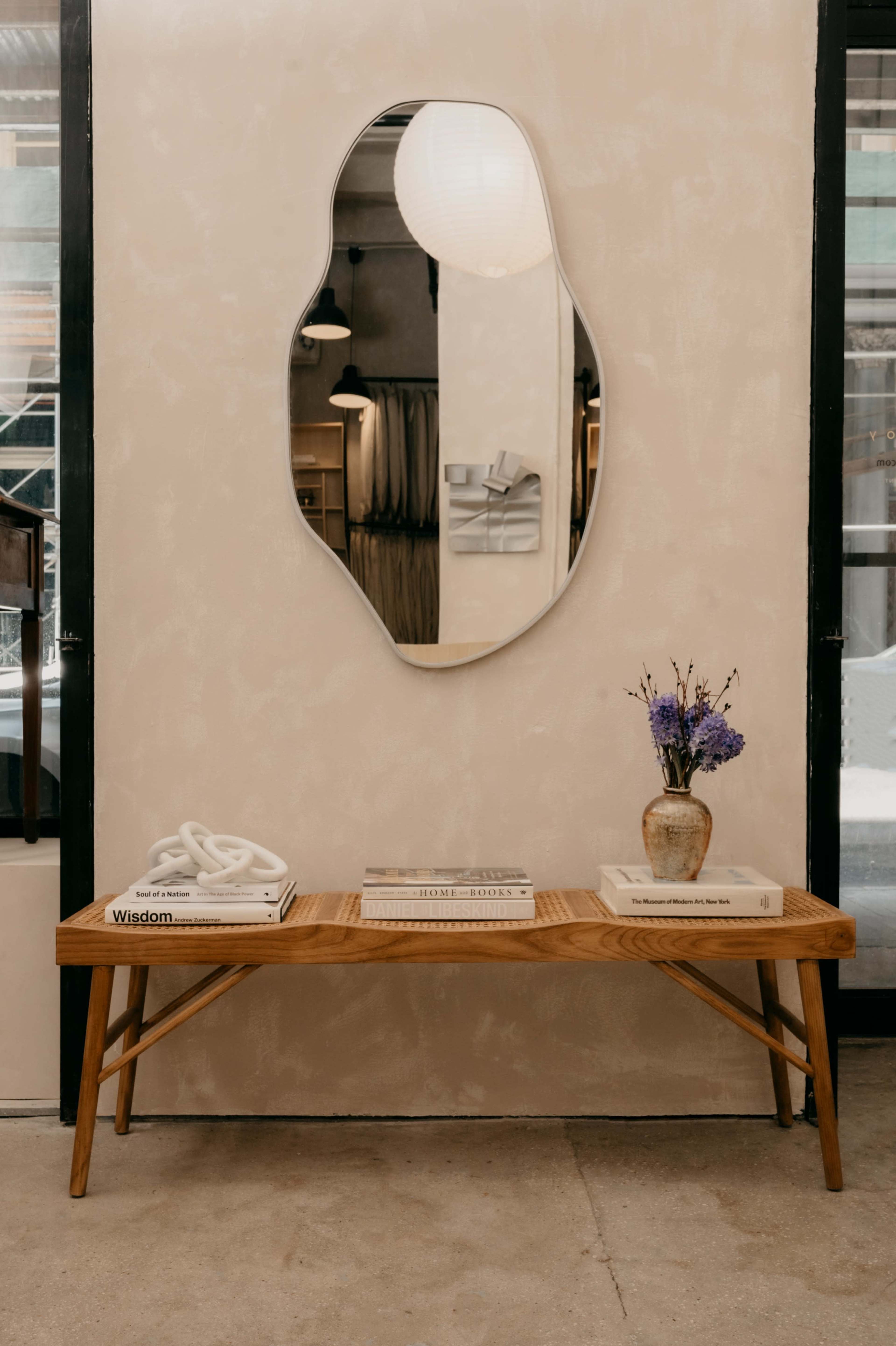A wooden bench with decorative books and a vase of flowers sits against a textured wall featuring a wavy mirror.