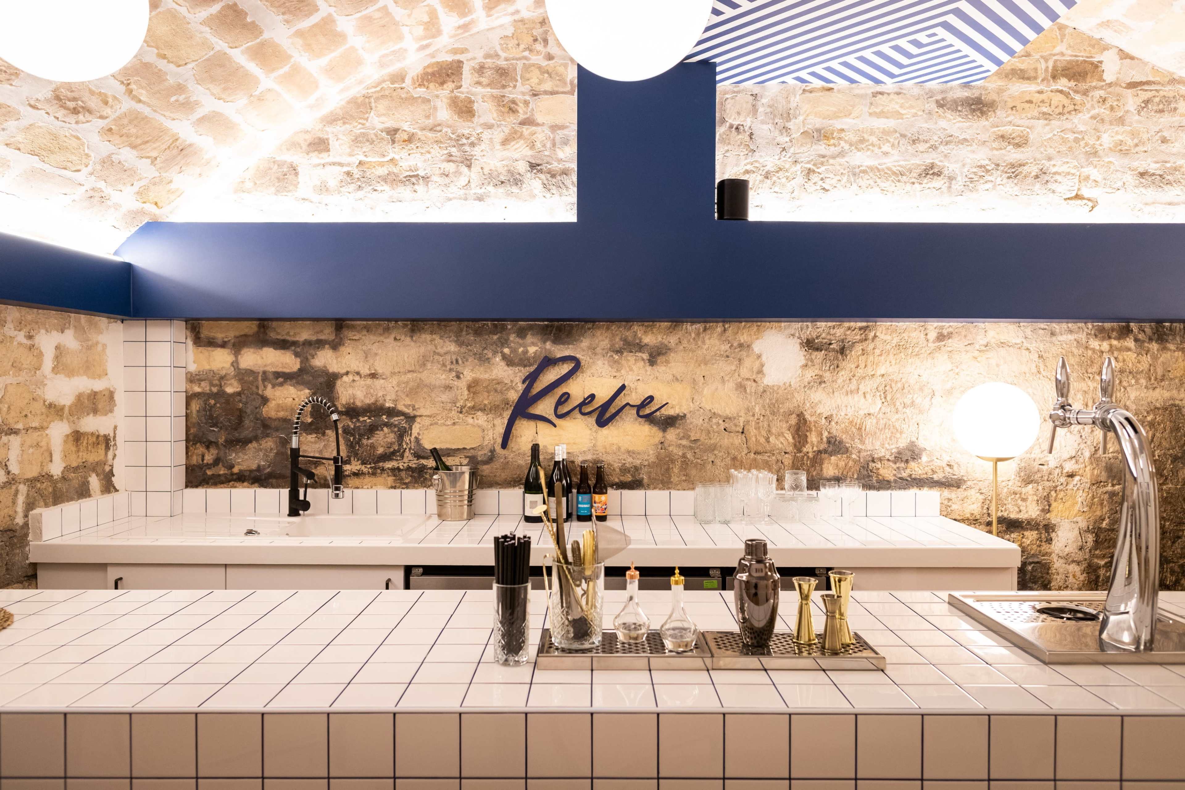 The image shows a modern bar with a white tiled countertop, featuring various barware and a blue accent wall with the word "Reeve" displayed.