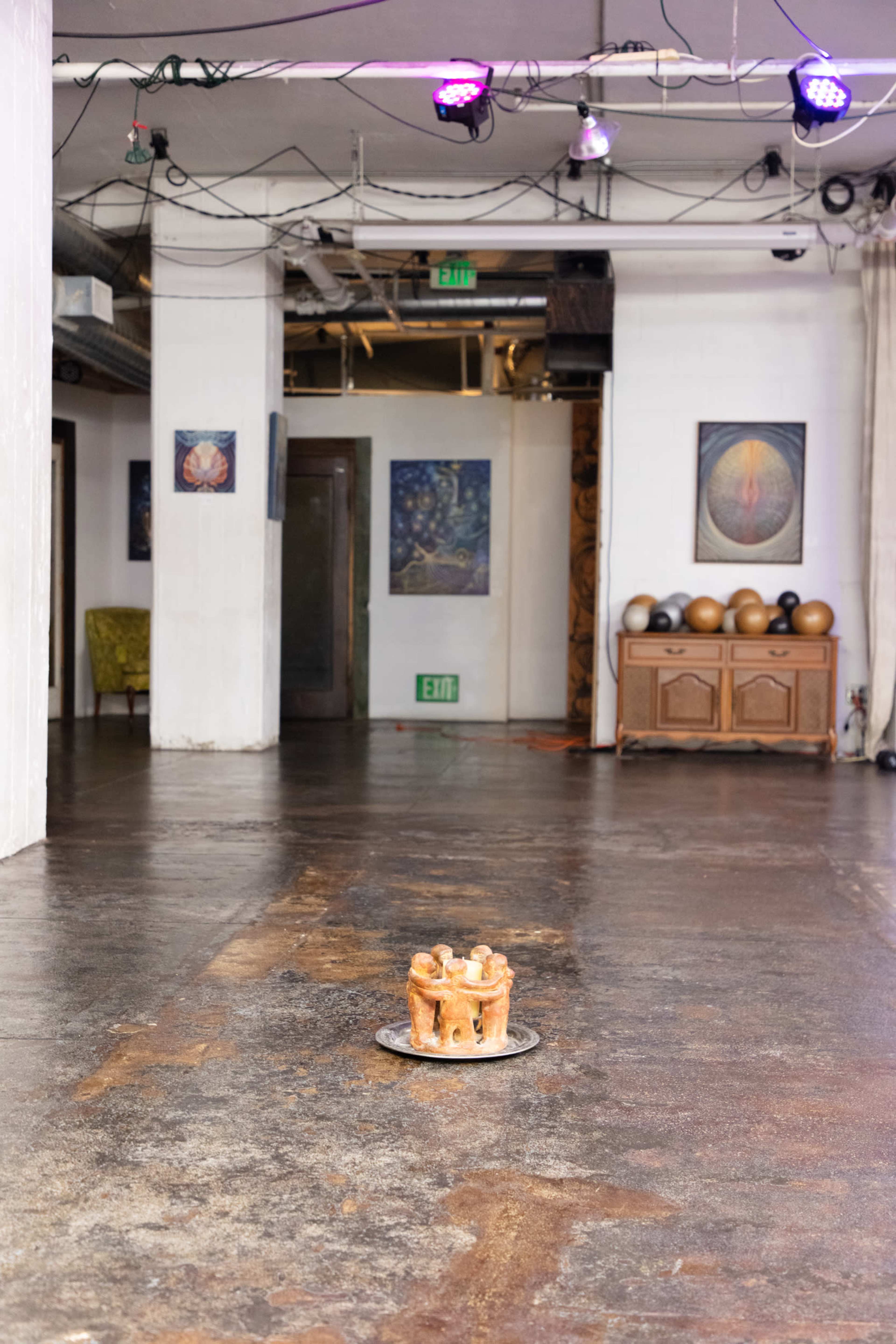 The image shows a sparse art studio with a small sculpture on a circular base in the center of a wooden floor, surrounded by walls adorned with framed artworks.