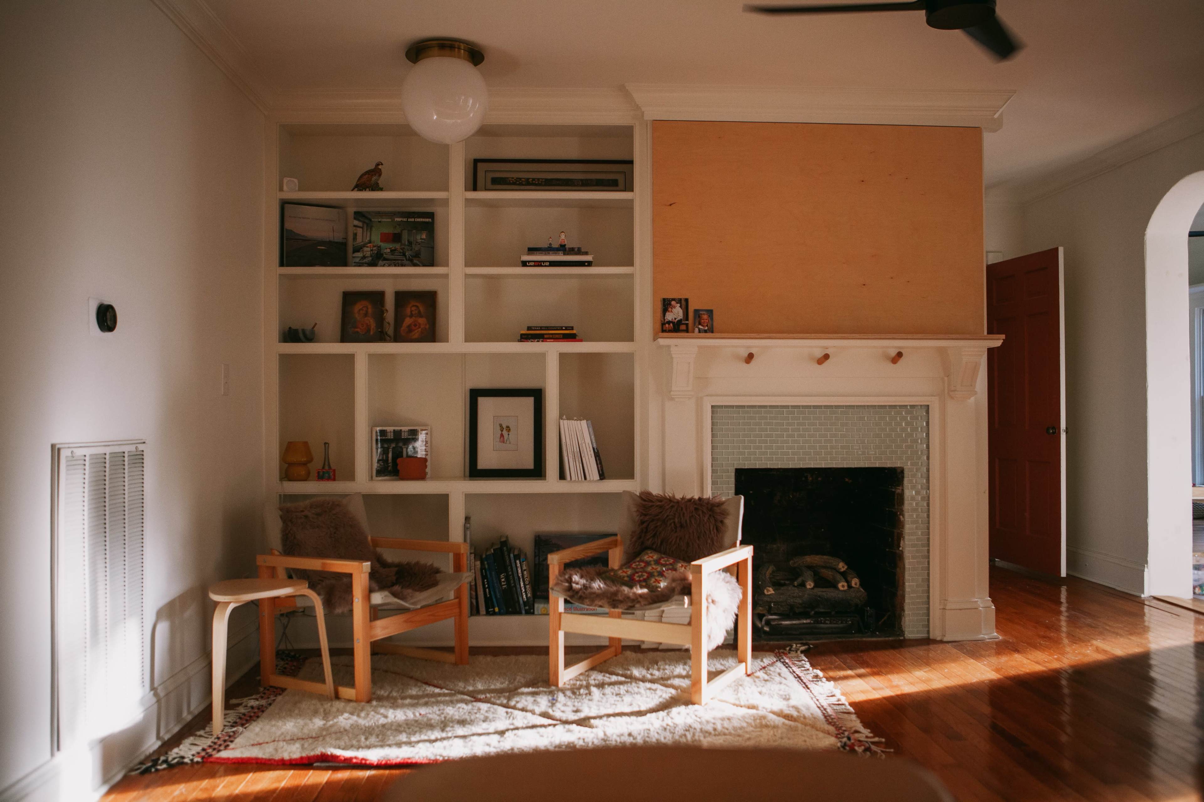 The image shows a living room featuring two wooden chairs with cushions, a white bookshelf filled with various items, and a fireplace on a wooden floor.
