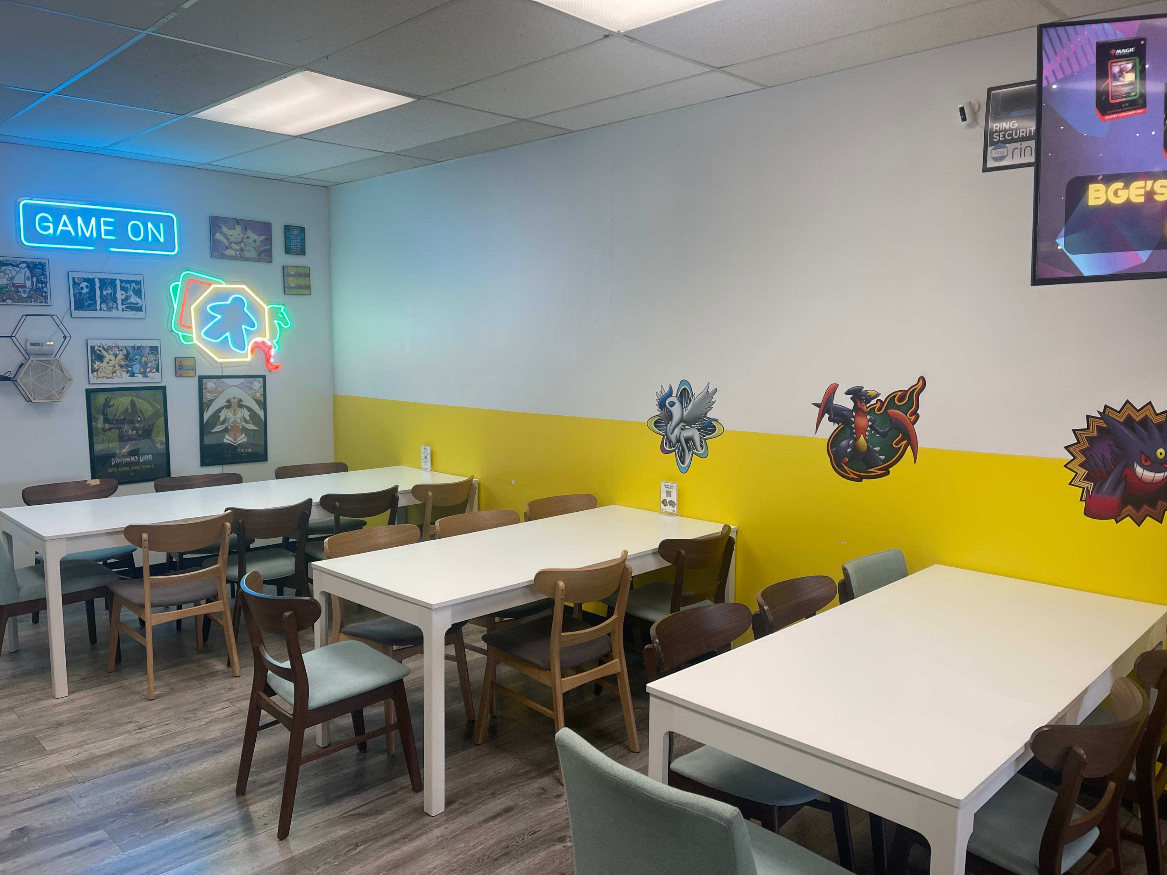 A brightly colored game room with white tables, wooden chairs, and wall decorations featuring characters and a neon sign that reads "GAME ON."