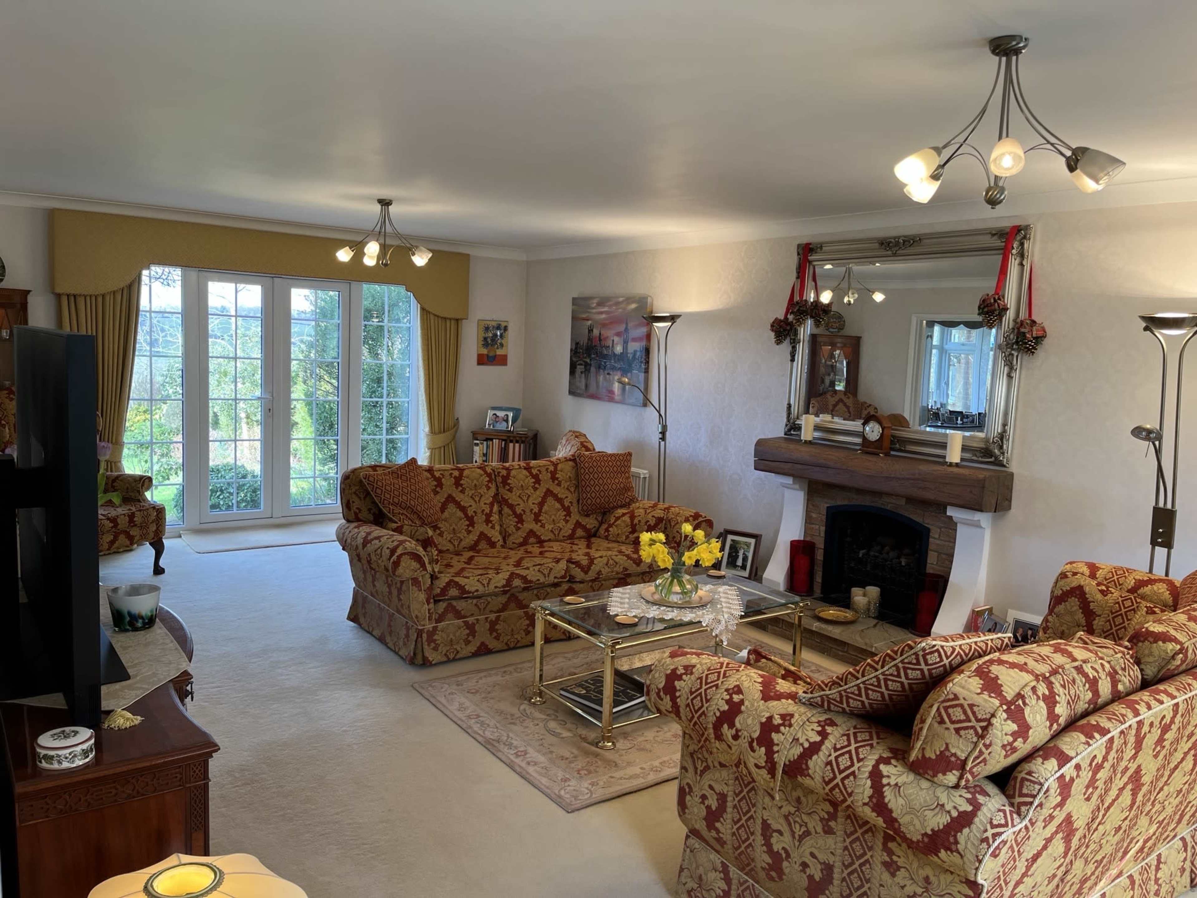 The living room features two patterned sofas, a glass coffee table, a fireplace with a mirror above it, and large windows allowing natural light.
