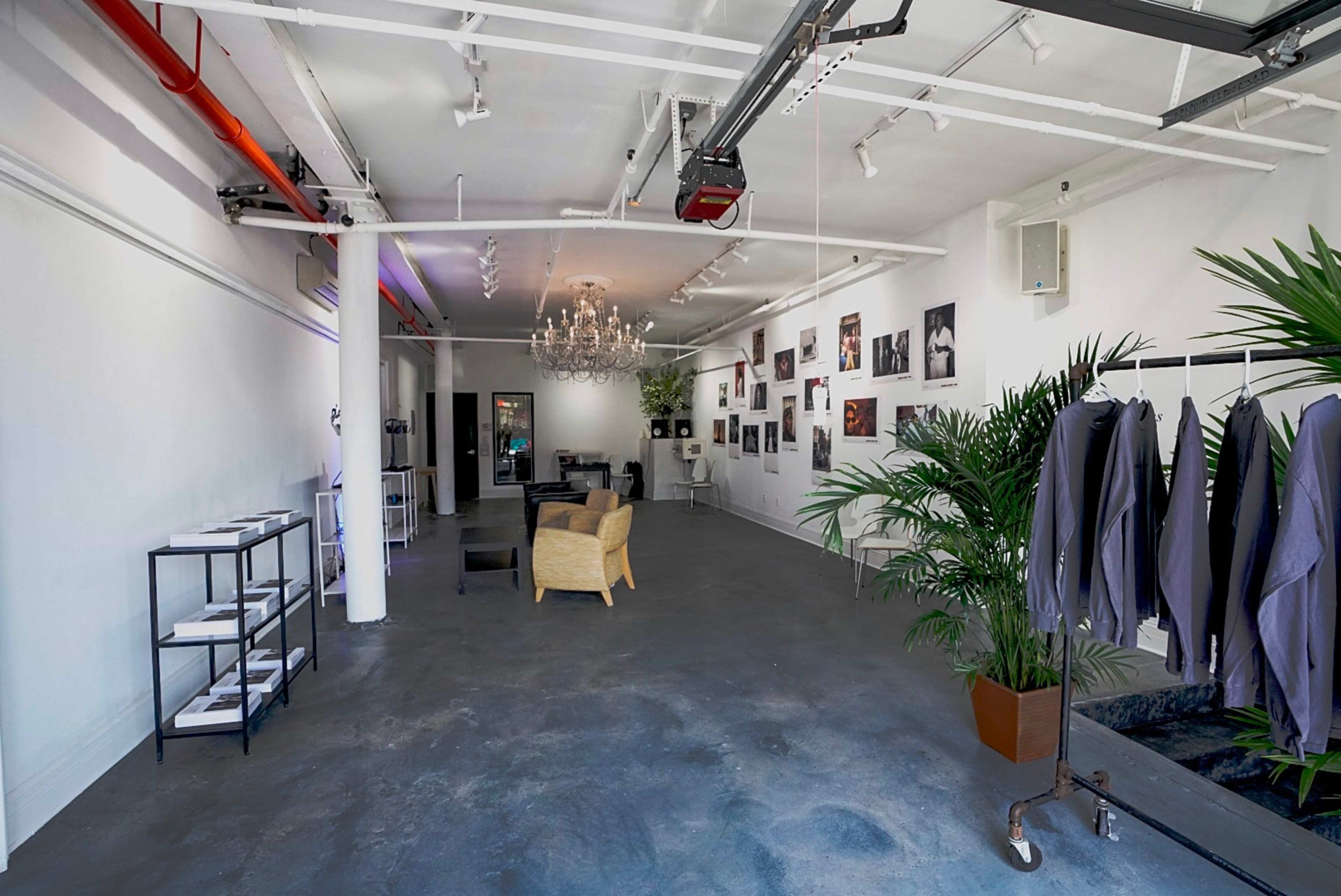 A spacious, well-lit gallery or studio with a chandelier, seating area, and clothing displayed on racks along one wall.
