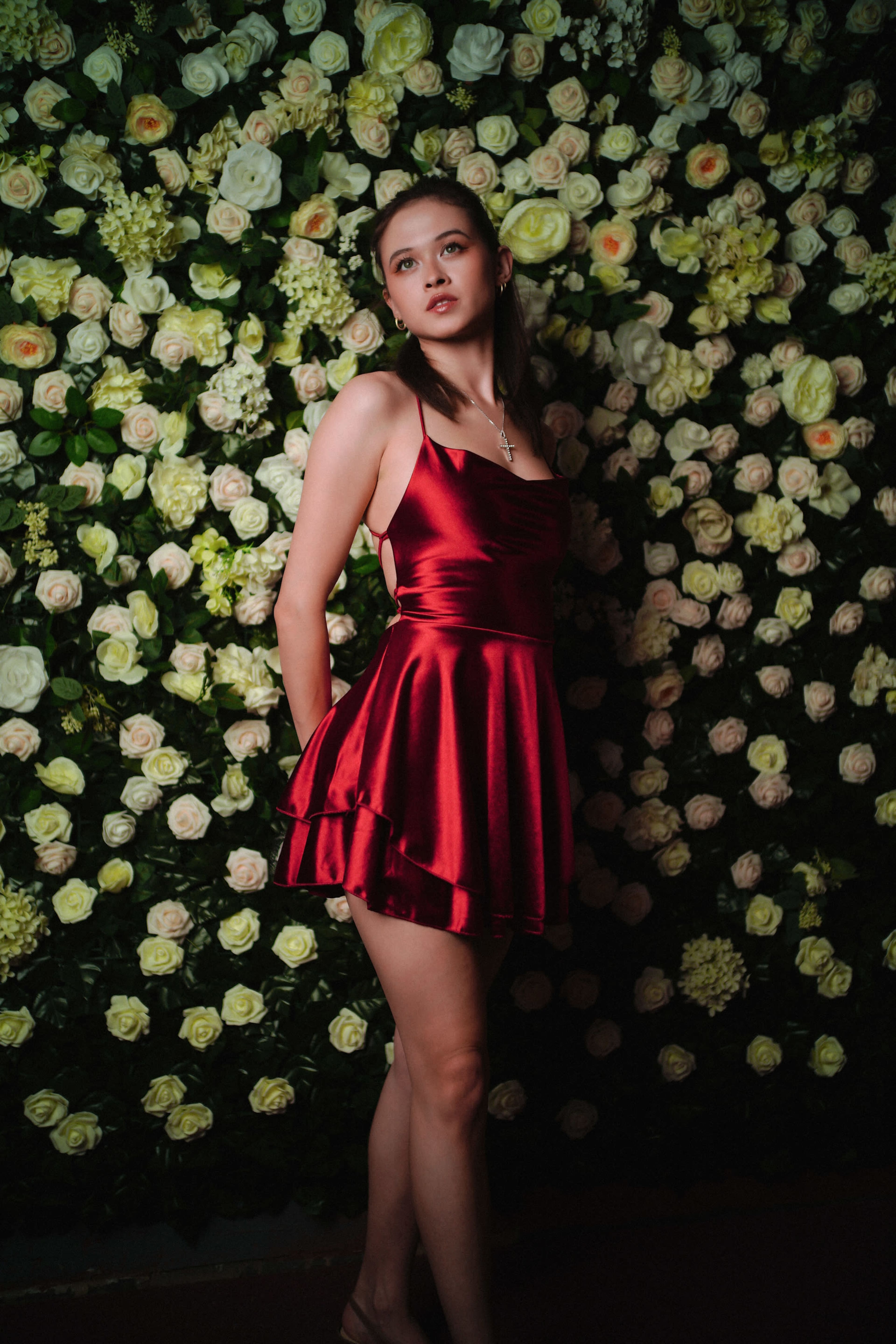A young woman in a red dress stands against a wall covered in white roses and greenery.