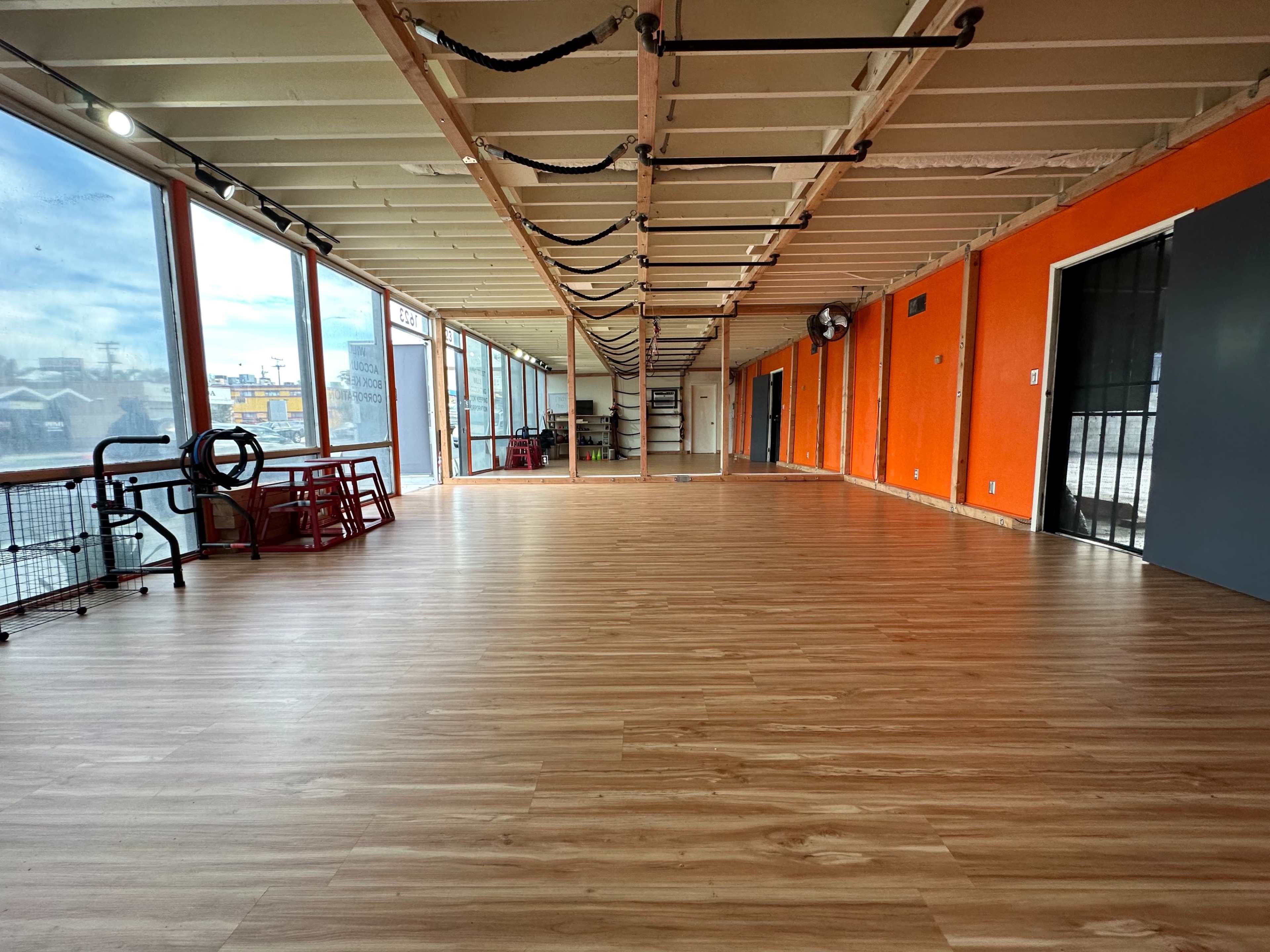 A spacious indoor area with wooden floors, large windows on one side, and orange walls, featuring empty space and some equipment in the background.