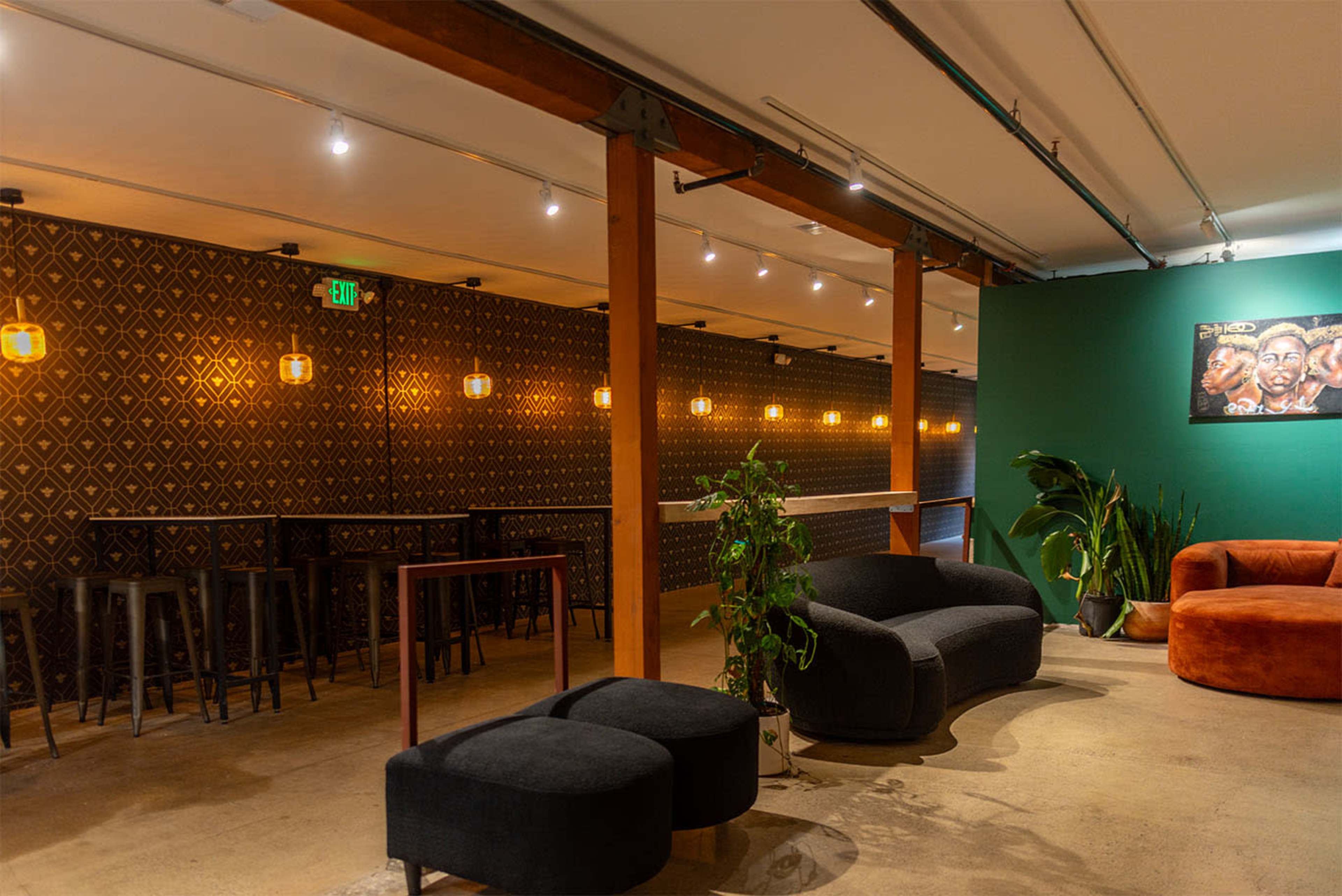 The image shows a modern, industrial-style lounge with a mix of seating options, including dark sofas and orange chairs, alongside a bar area and decorative plants.