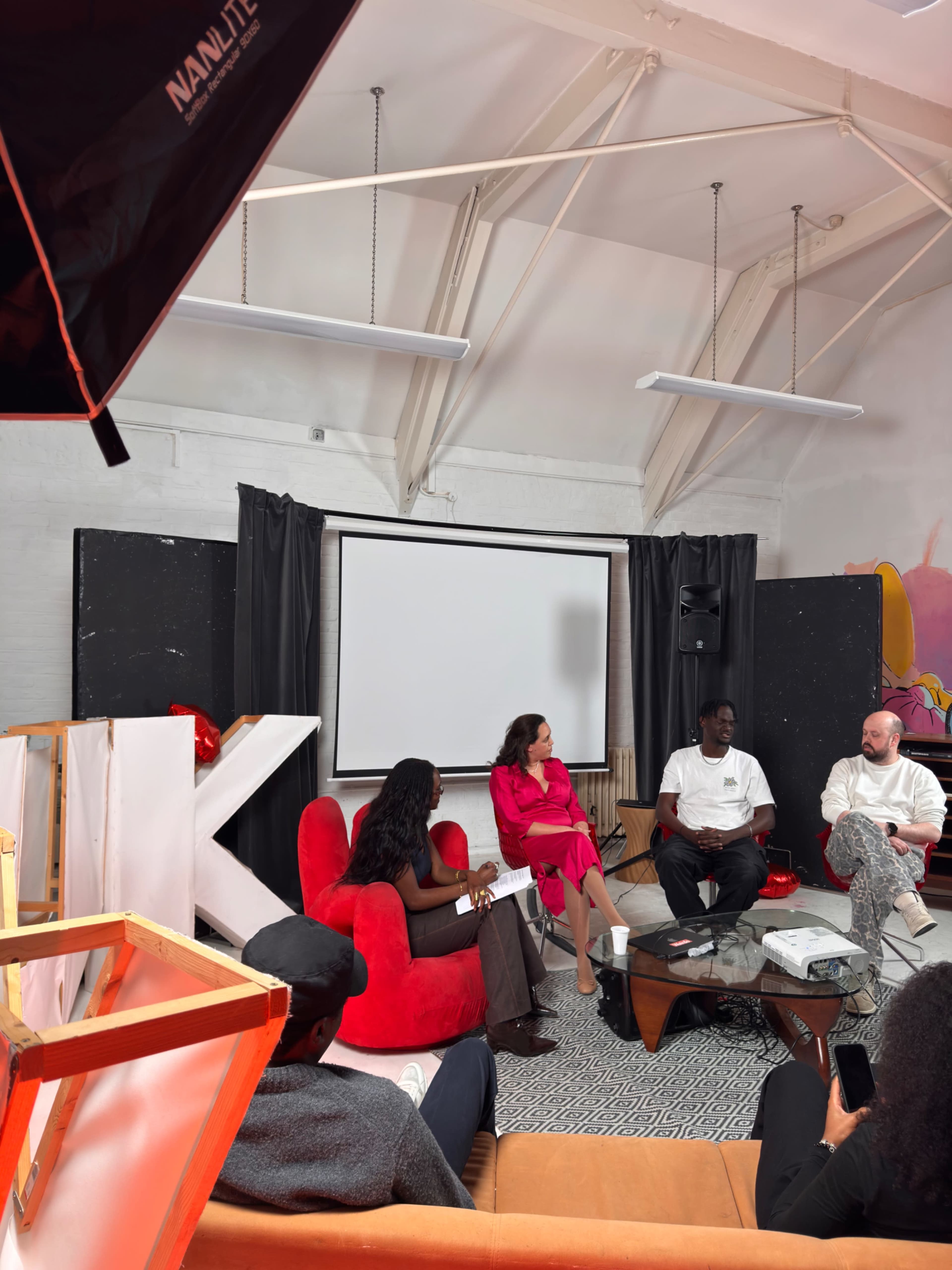A panel discussion takes place in a creative workspace, featuring three speakers seated on chairs, with an audience taking notes.