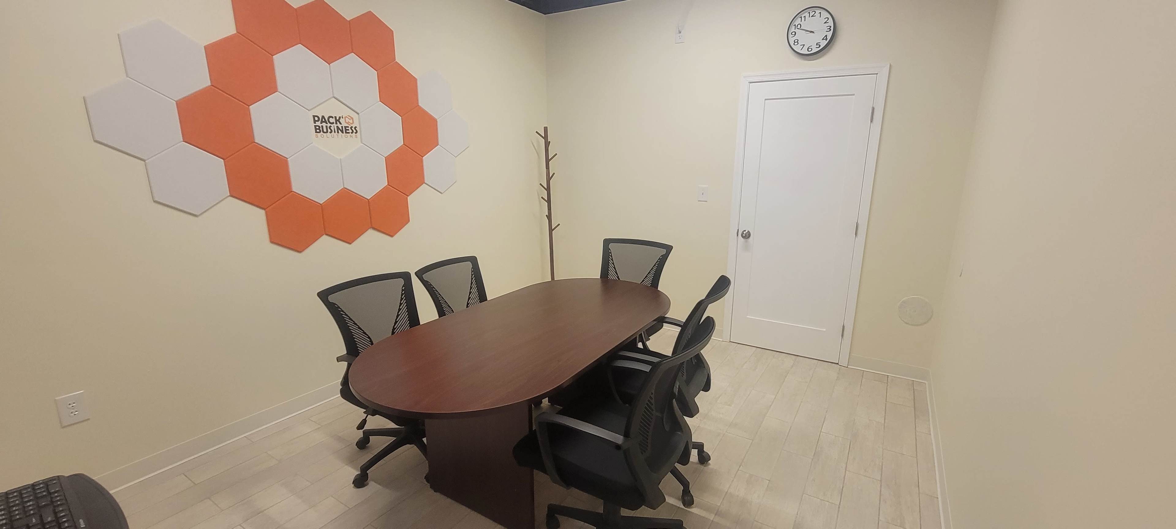 A small conference room with a long table surrounded by six black chairs, a clock on the wall, and hexagonal orange and white wall decor.
