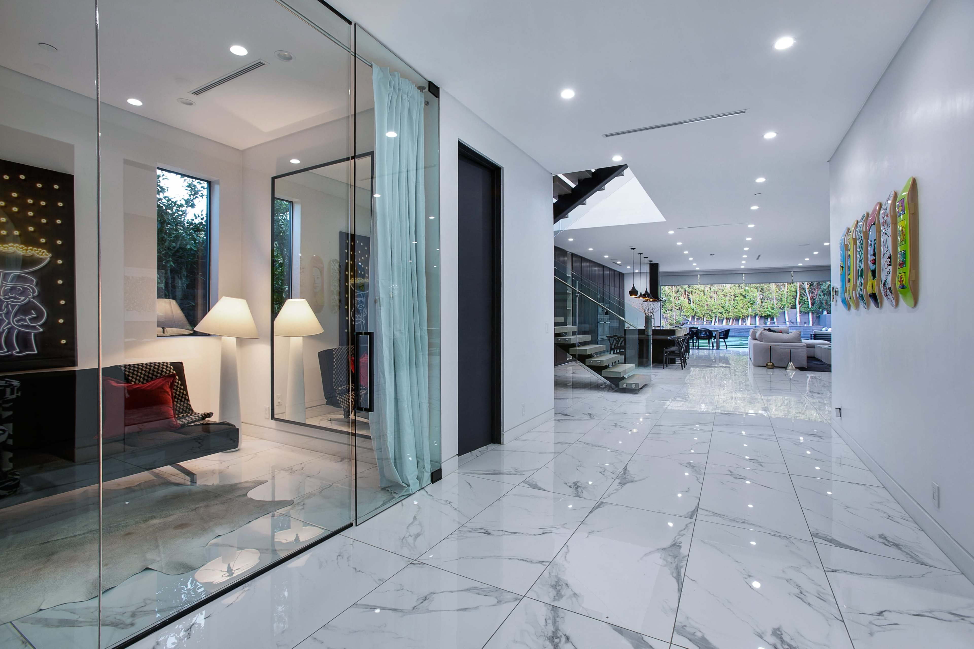 The image shows a modern hallway with marble flooring, glass walls, and a view into an adjoining living space featuring stairs and large windows.