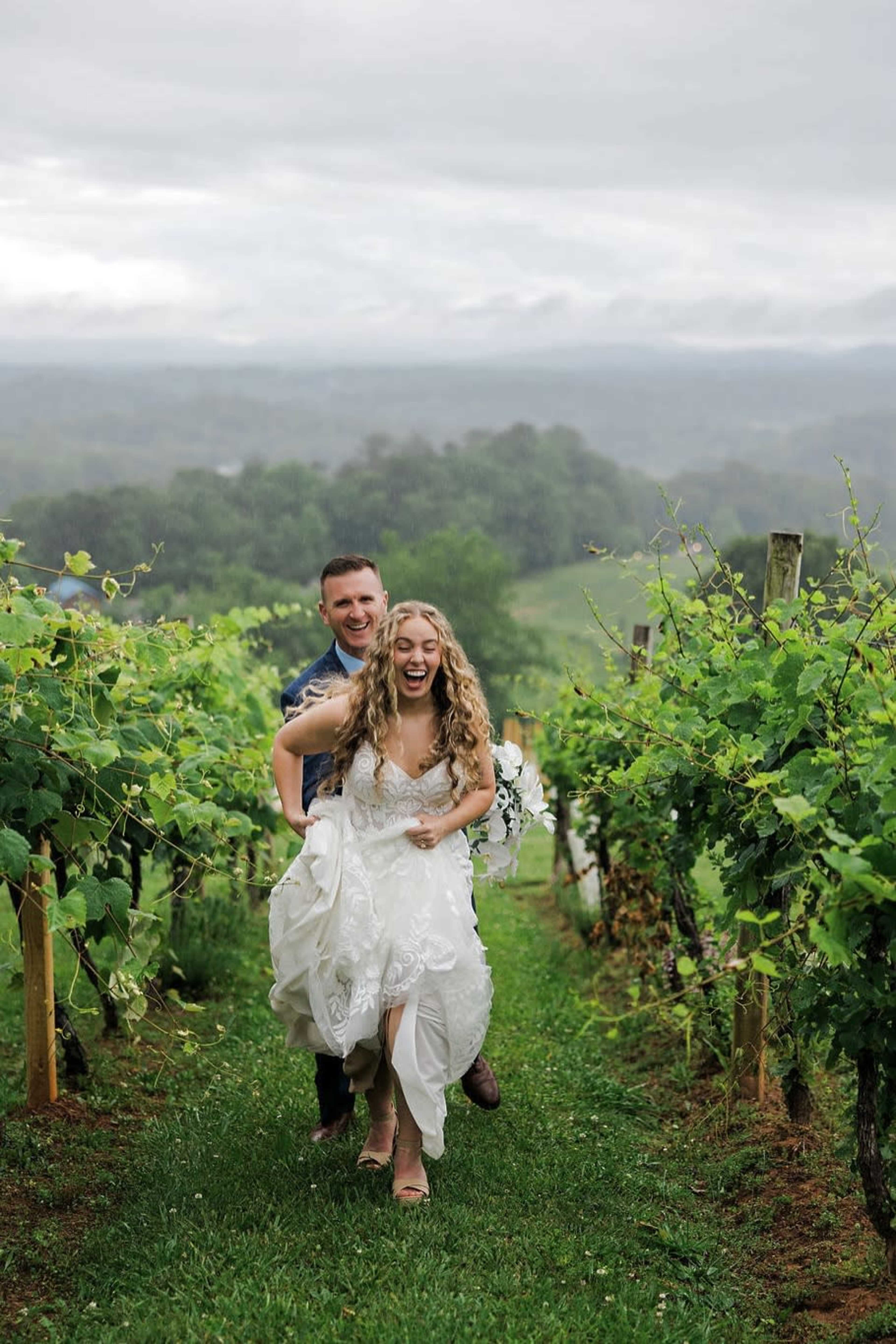 A bride and groom joyfully run together through a vineyard on a cloudy day.