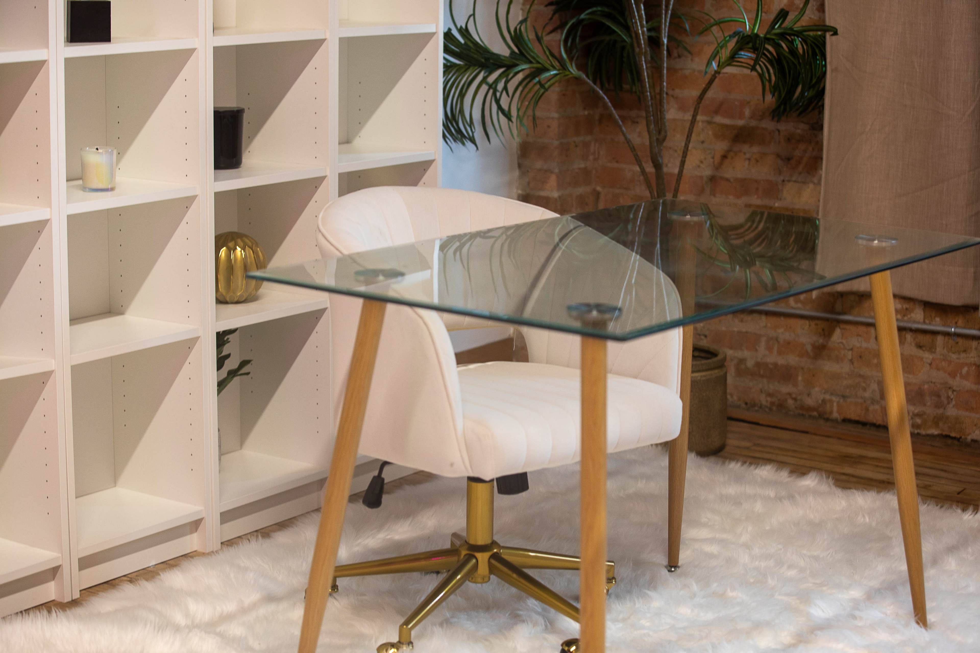 A modern workspace features a glass-top desk, a white upholstered chair, and a bookshelf against a brick wall.