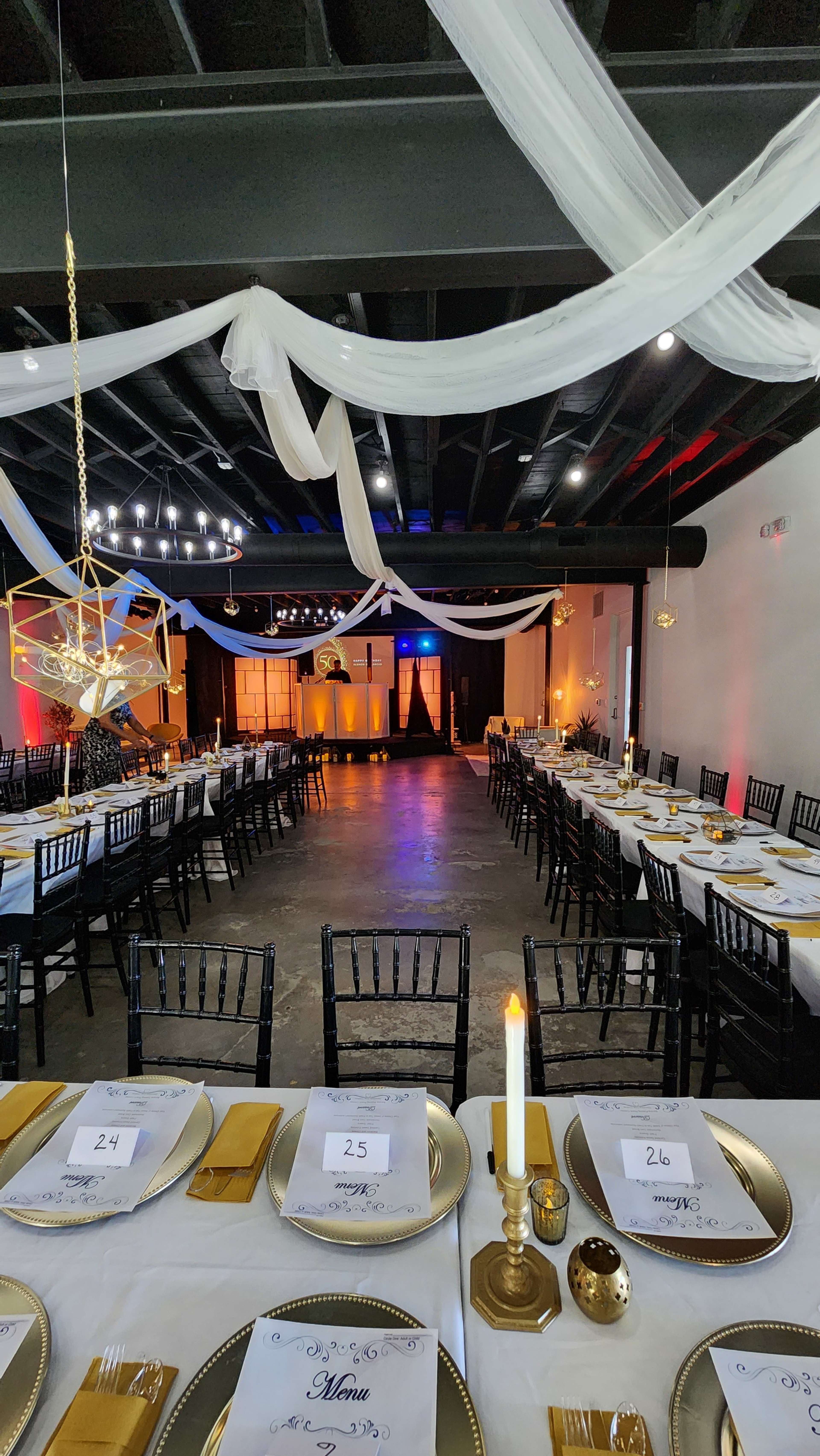 The image shows a banquet hall set up for an event, featuring long tables adorned with gold tableware, white menus, and decorative lighting, with a stage in the background.