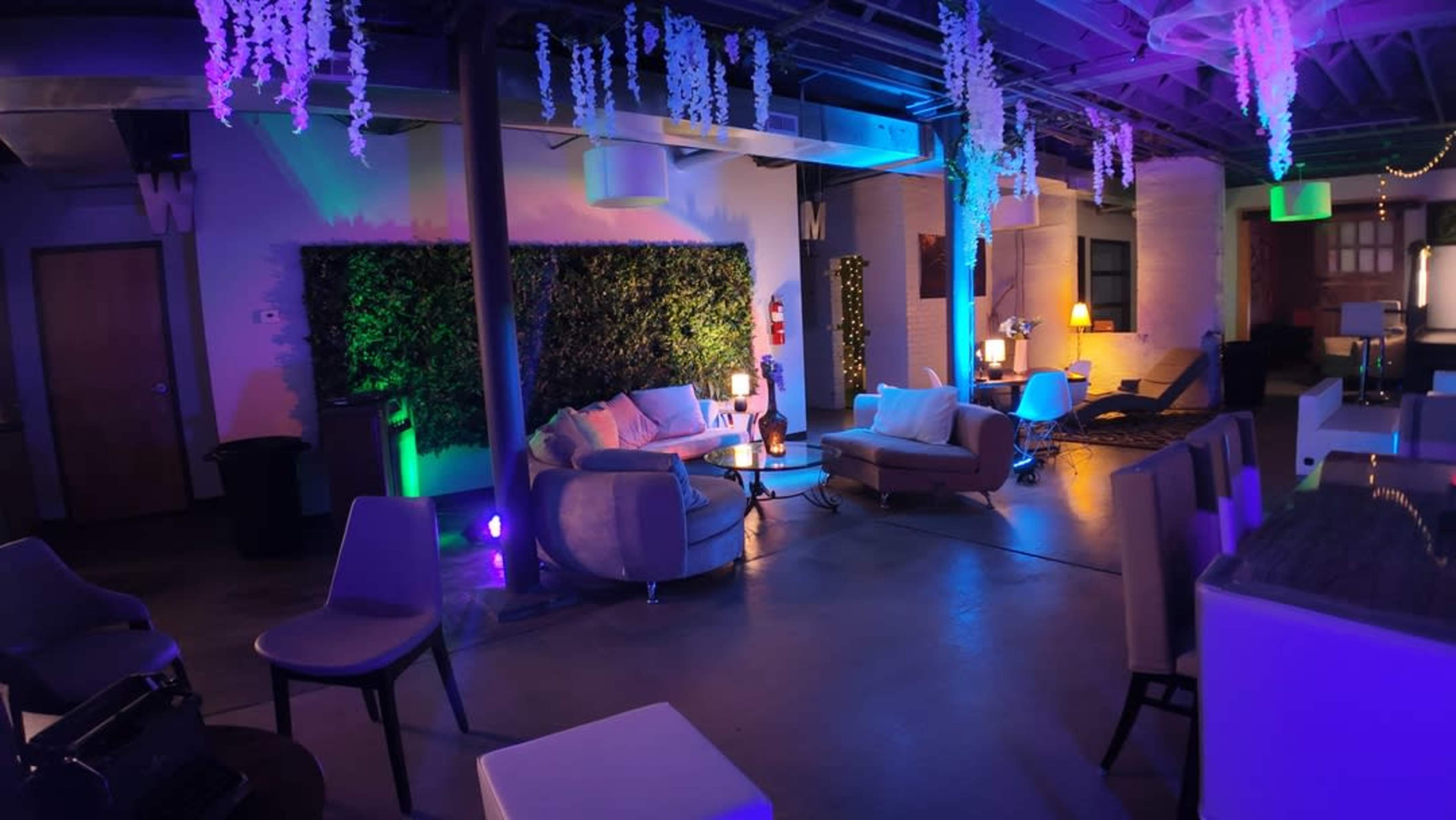The image shows a dimly lit lounge area with modern furniture, decorative plants on the walls, and colorful lighting fixtures.
