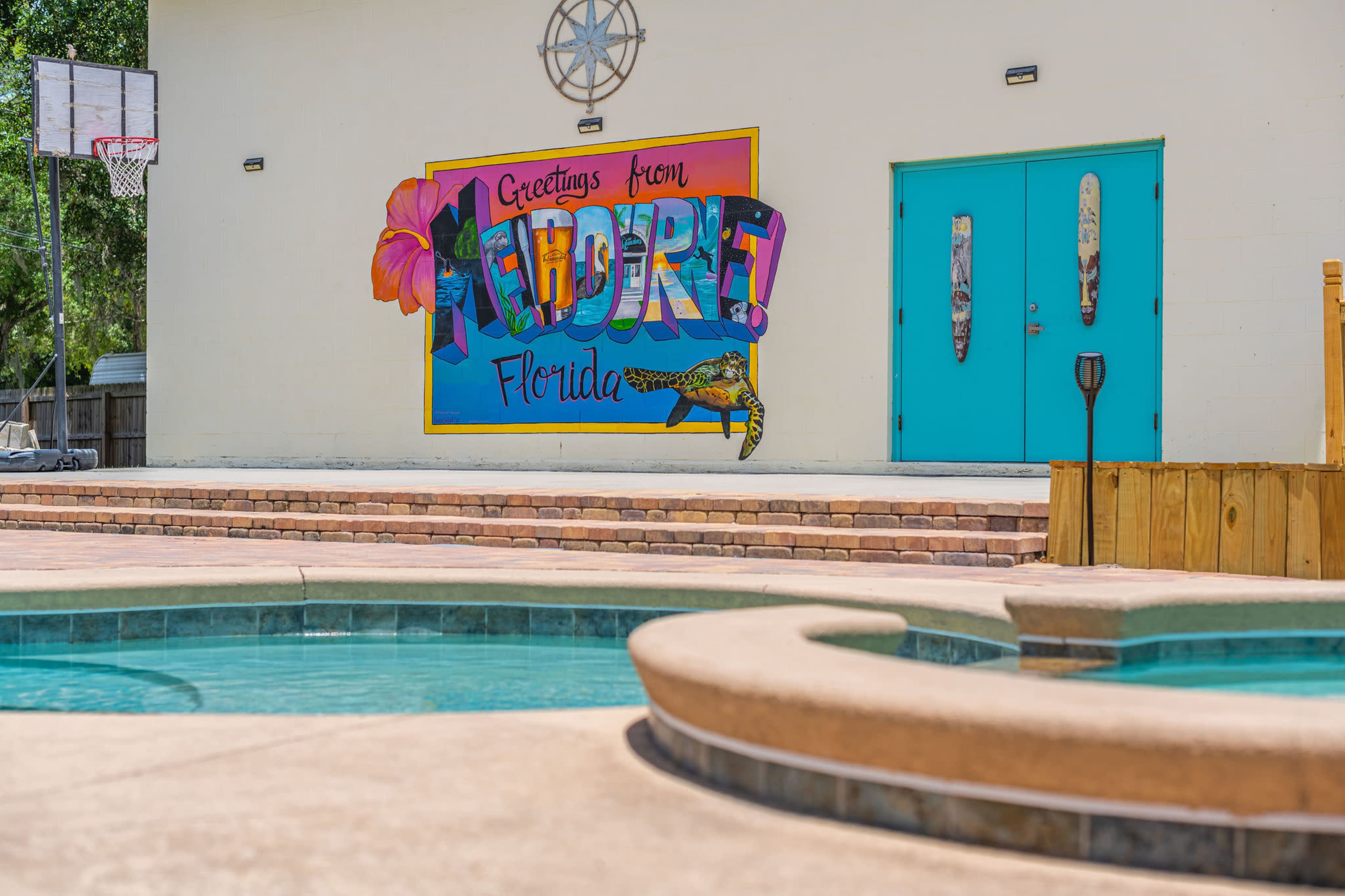 A colorful mural reading "Greetings from Florida" is displayed on a wall near a swimming pool area with a hot tub, basketball hoop, and turquoise doors in the background.