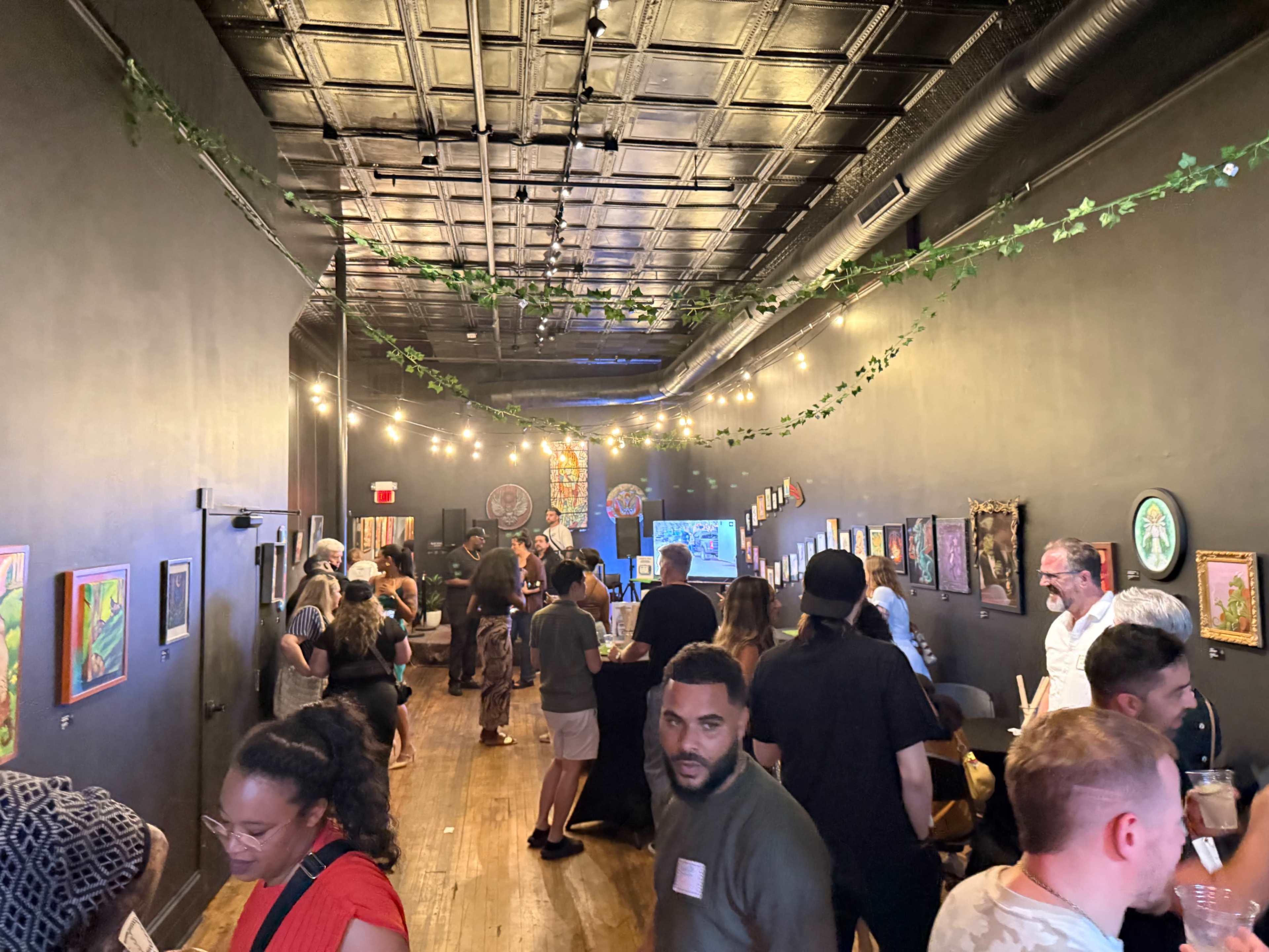 A crowded art gallery features people mingling and viewing various artworks while string lights illuminate the space.
