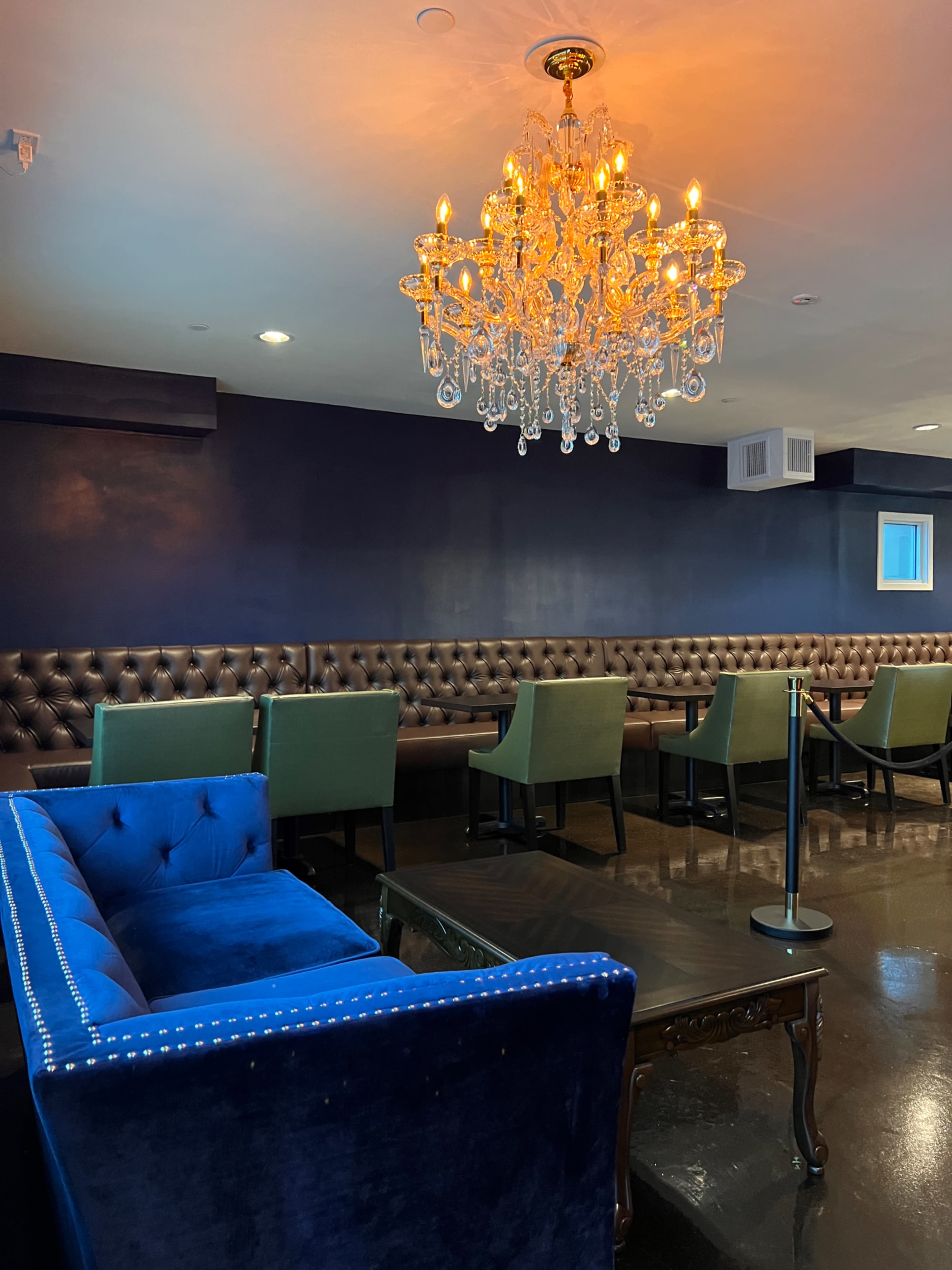 The image shows a lounge area featuring a blue velvet sofa, a dark wooden coffee table, a row of green chairs, and a decorative chandelier overhead against a navy blue wall.