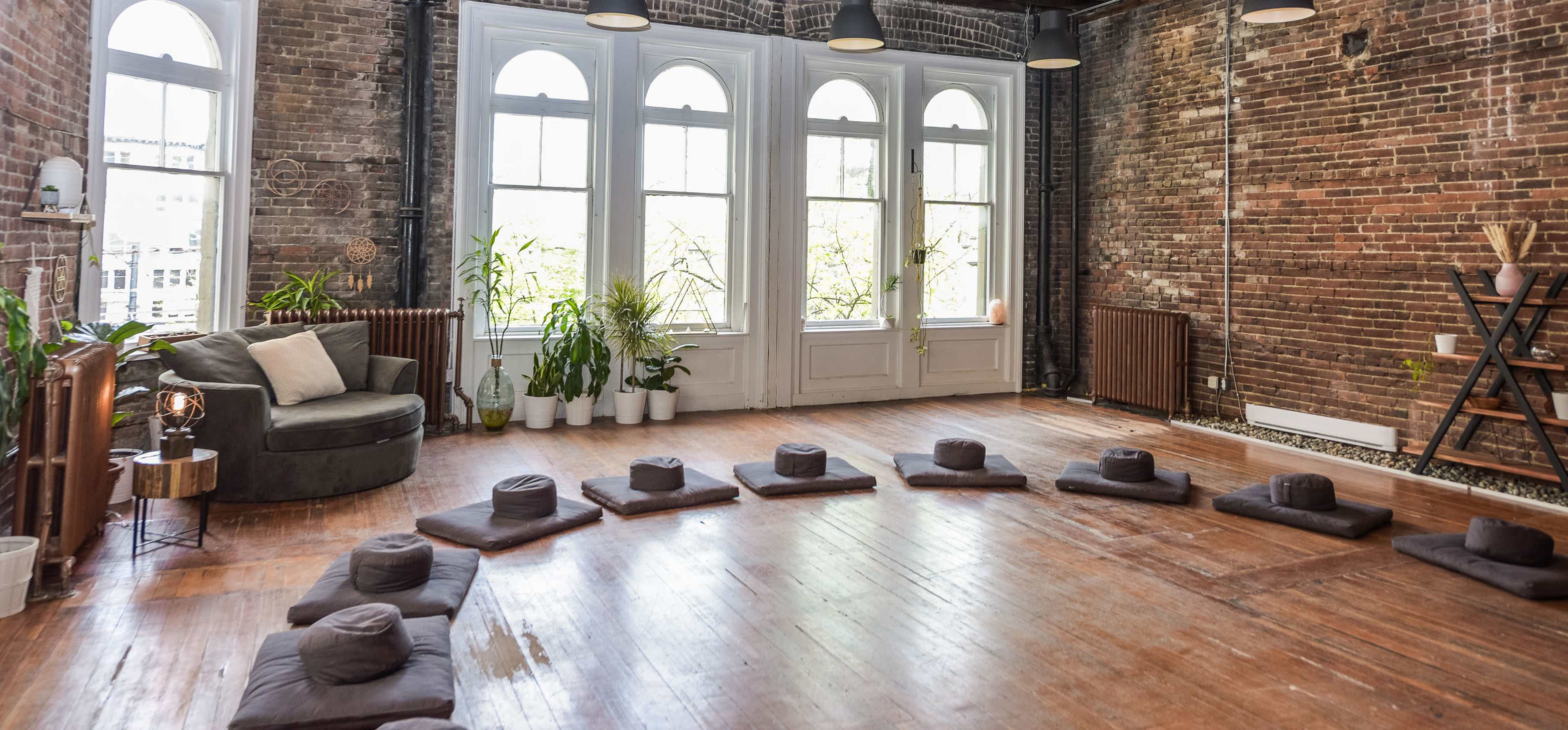 A spacious, minimalist room features a circular arrangement of meditation cushions on a wooden floor, surrounded by large windows and greenery.