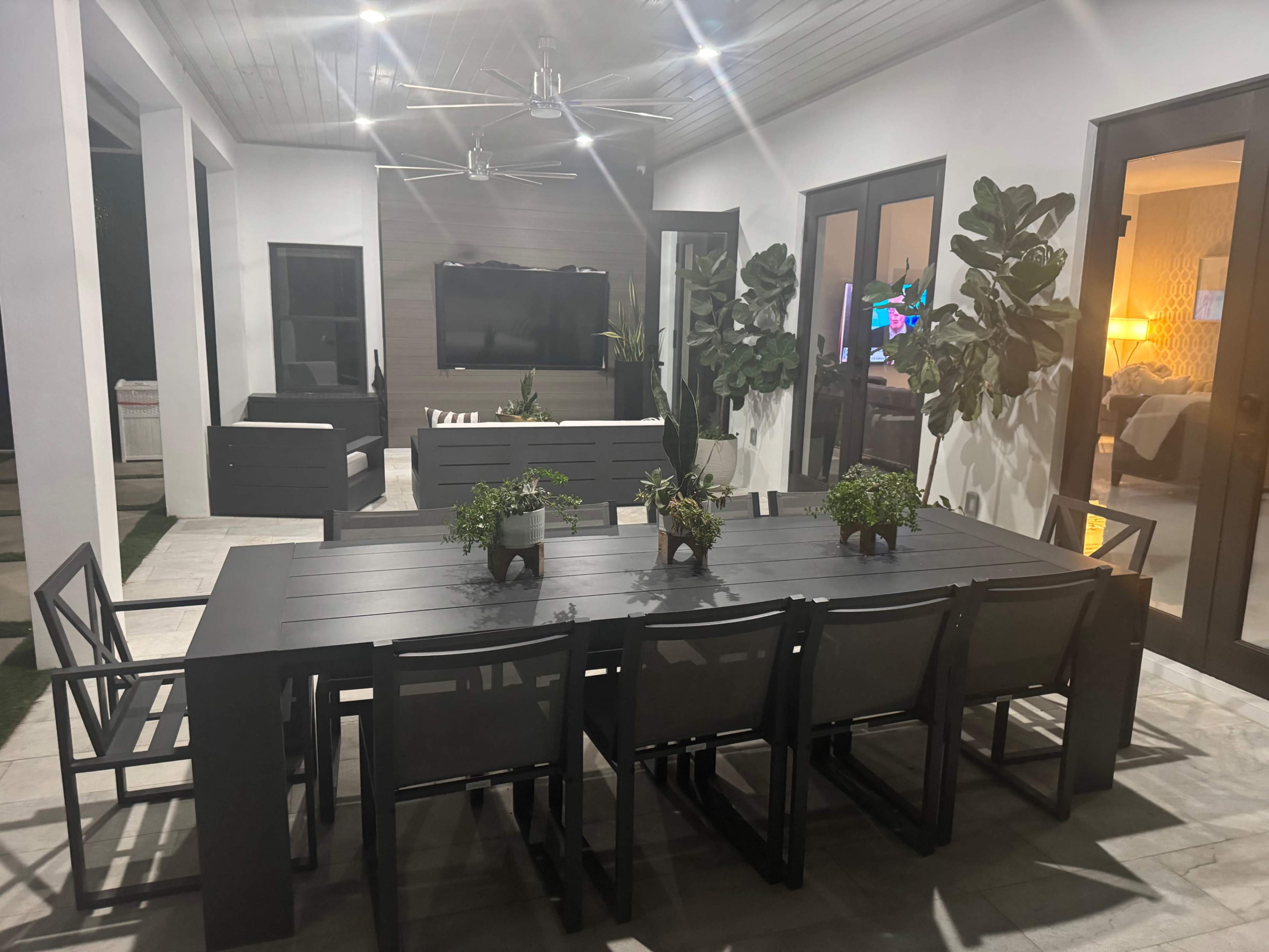 The image shows a modern outdoor dining area with a long table and several chairs, surrounded by potted plants and featuring a ceiling fan and a television on the wall.