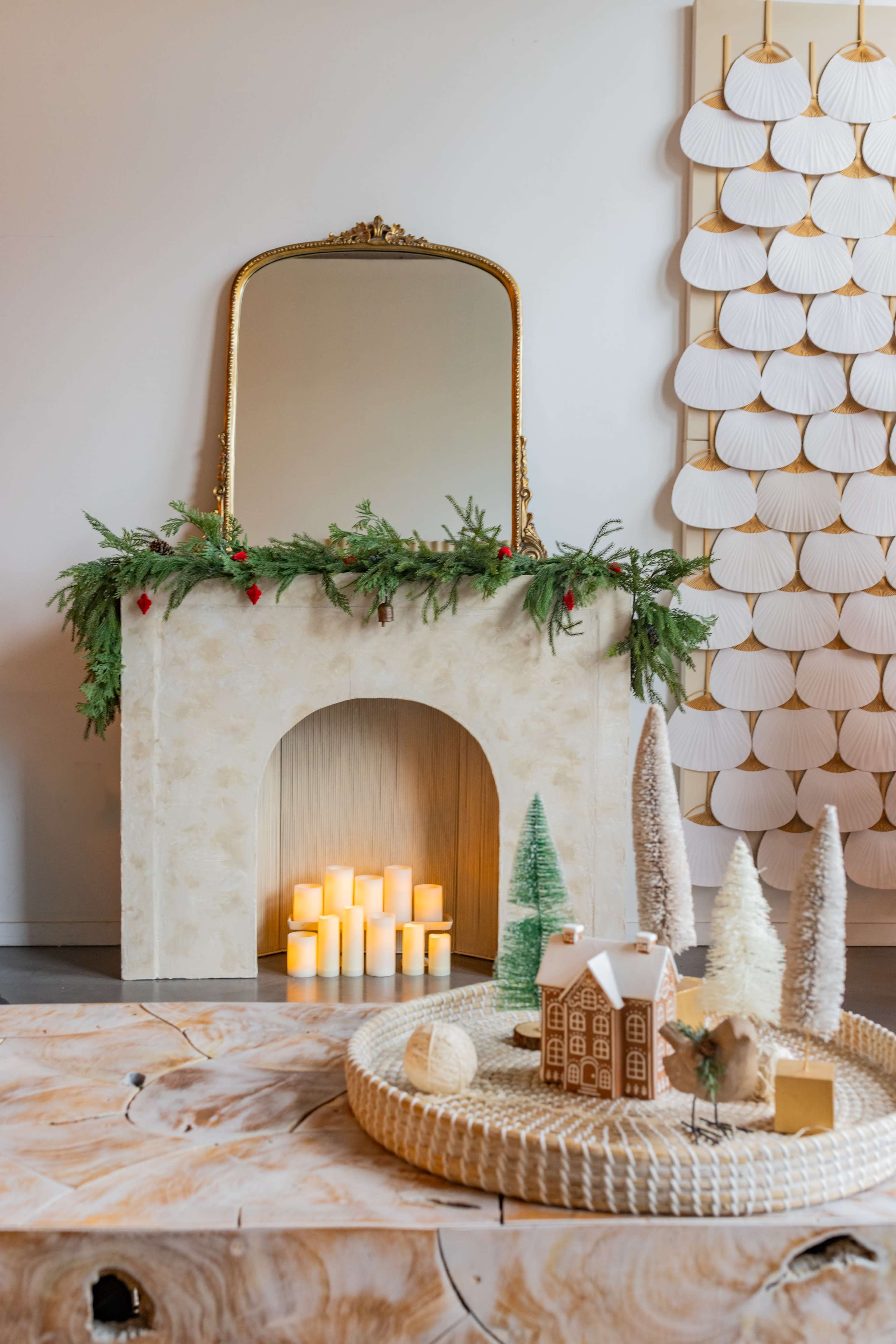 A decorated fireplace with a mirror above it, surrounded by greenery and candles, is positioned next to a wall adorned with light-colored fan-shaped decor.