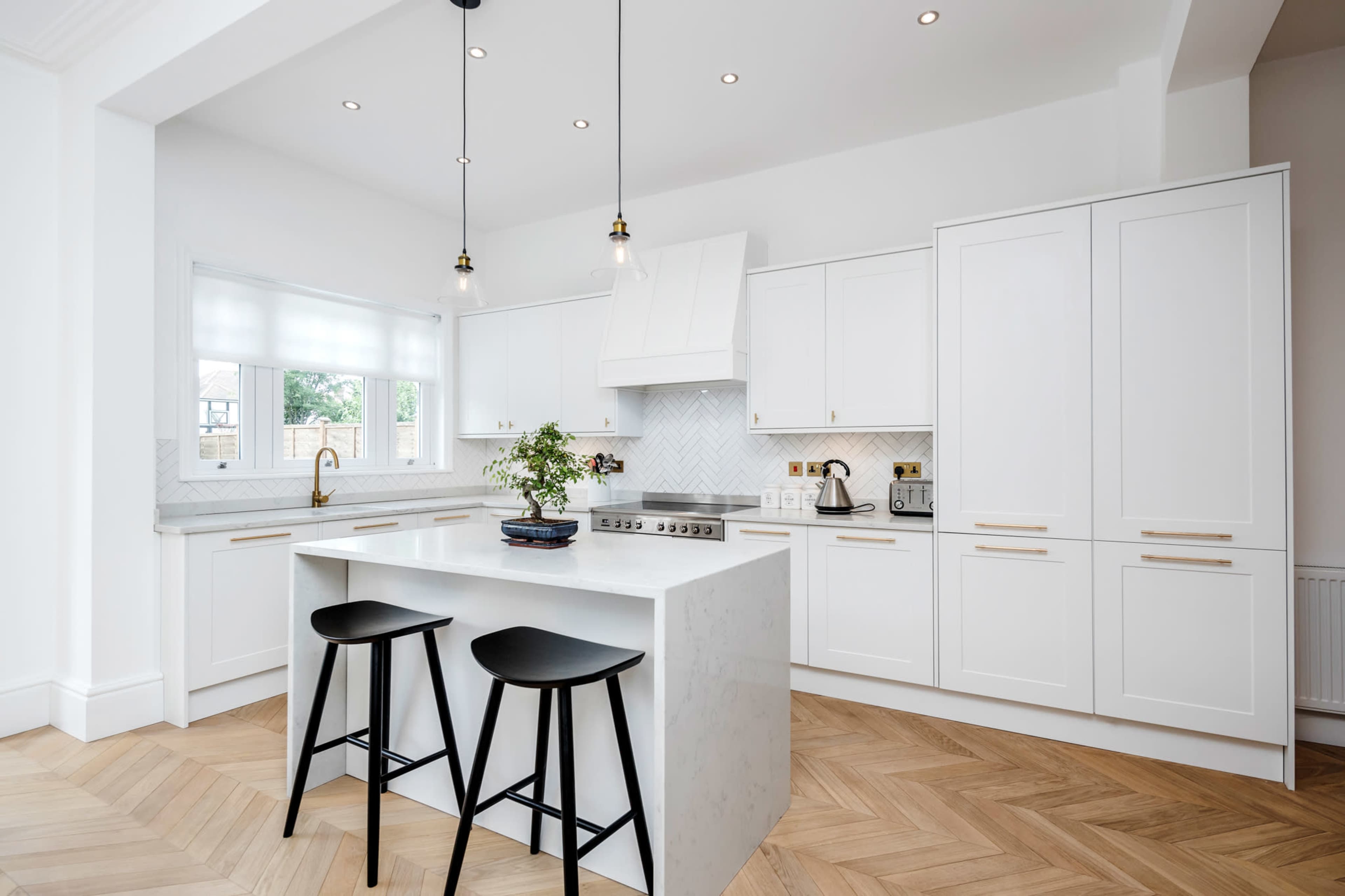 A modern kitchen features white cabinetry, a central island with two black stools, and herringbone-patterned wooden flooring.