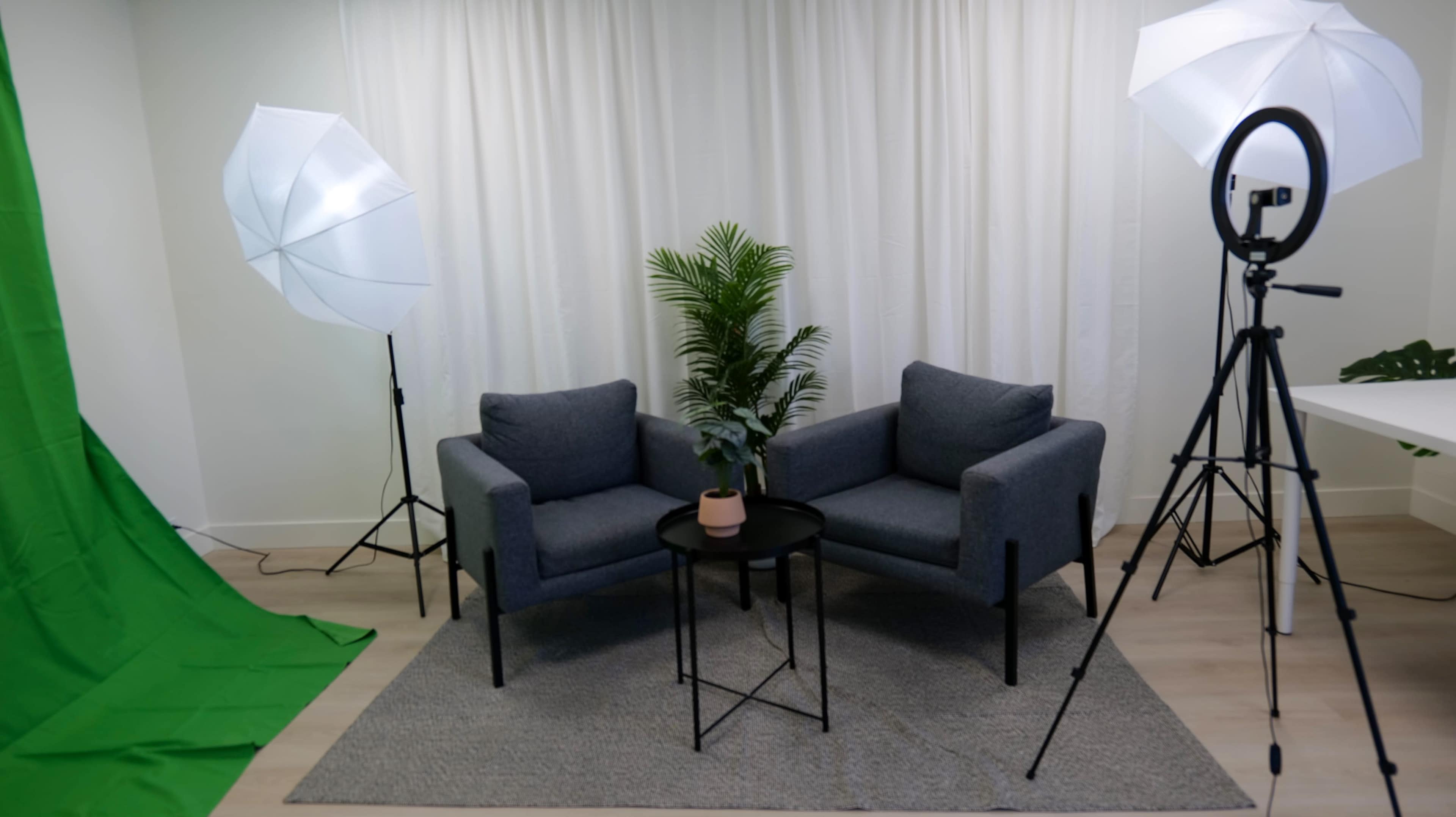 A simple studio setup with two grey chairs, a small black table, a potted plant, and lighting equipment, all against a backdrop of white curtains and a green screen.