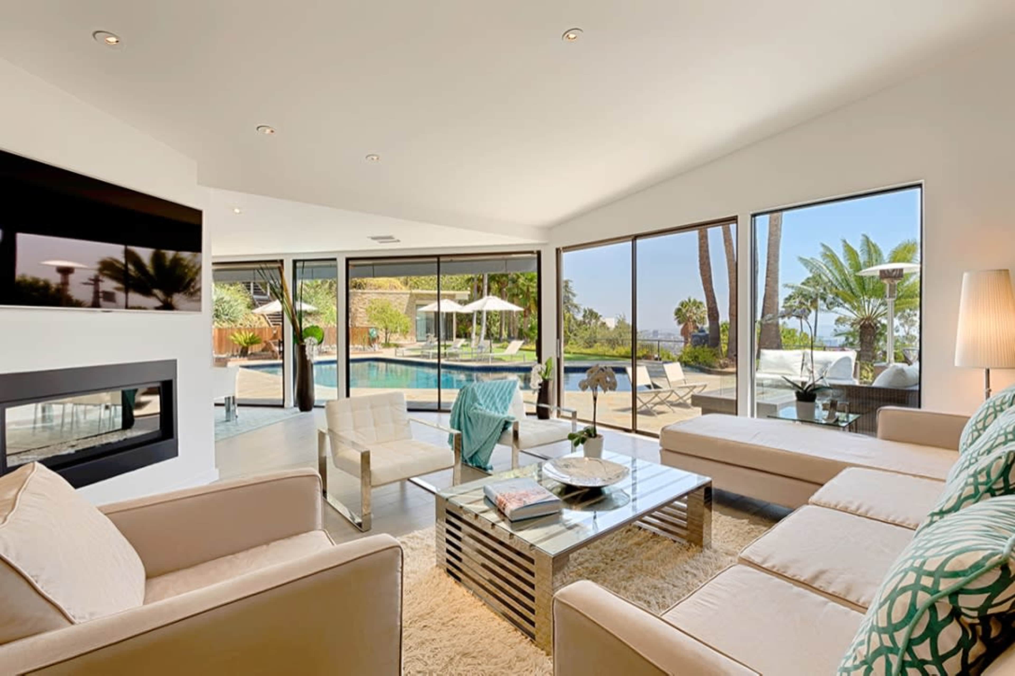 A modern living room features a beige sectional sofa, a glass coffee table, and large windows overlooking a pool and palm trees.