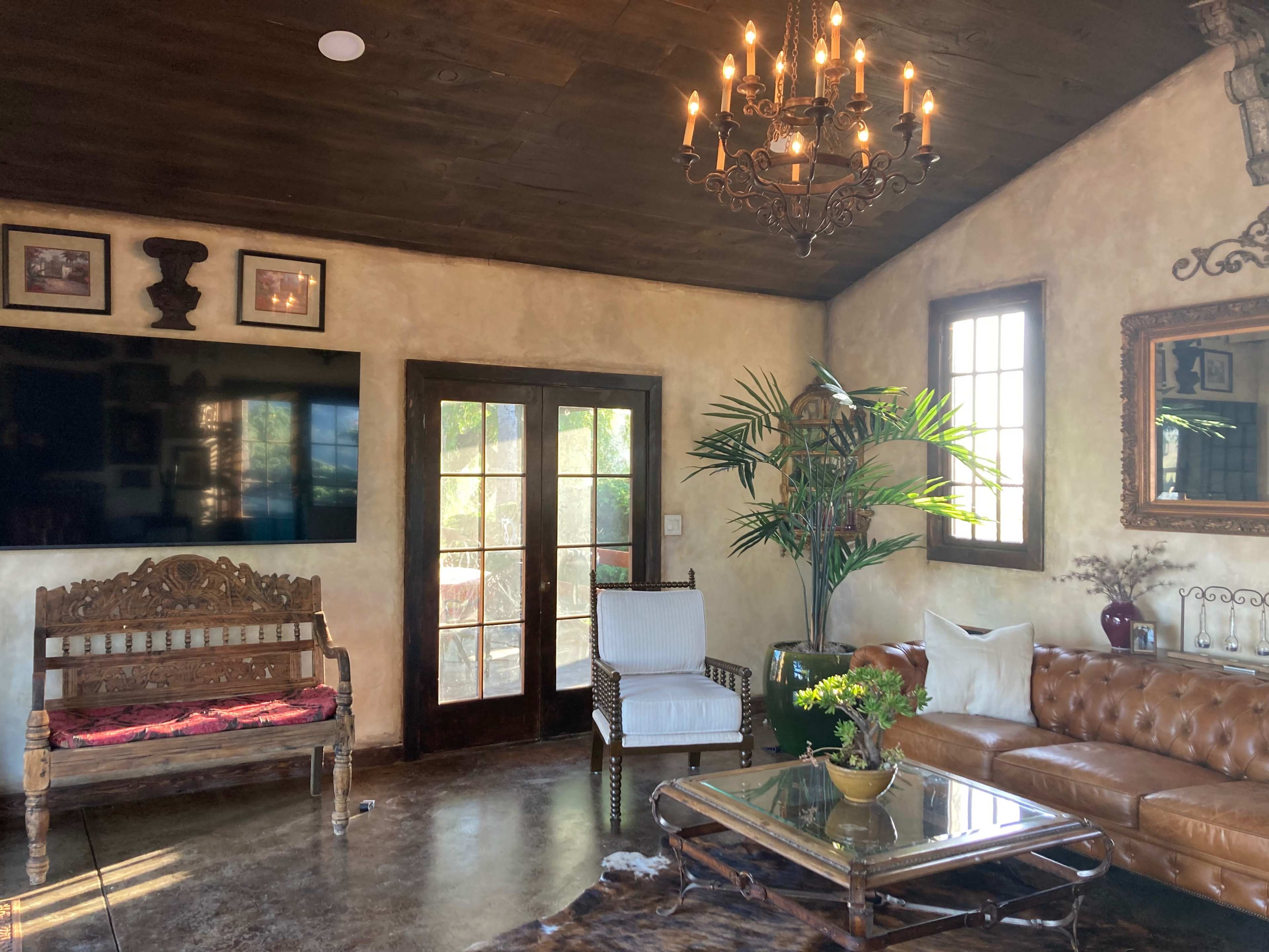 The image shows a living room with a chandelier, a wooden bench, a leather sofa, a glass coffee table, and large windows featuring greenery outside.