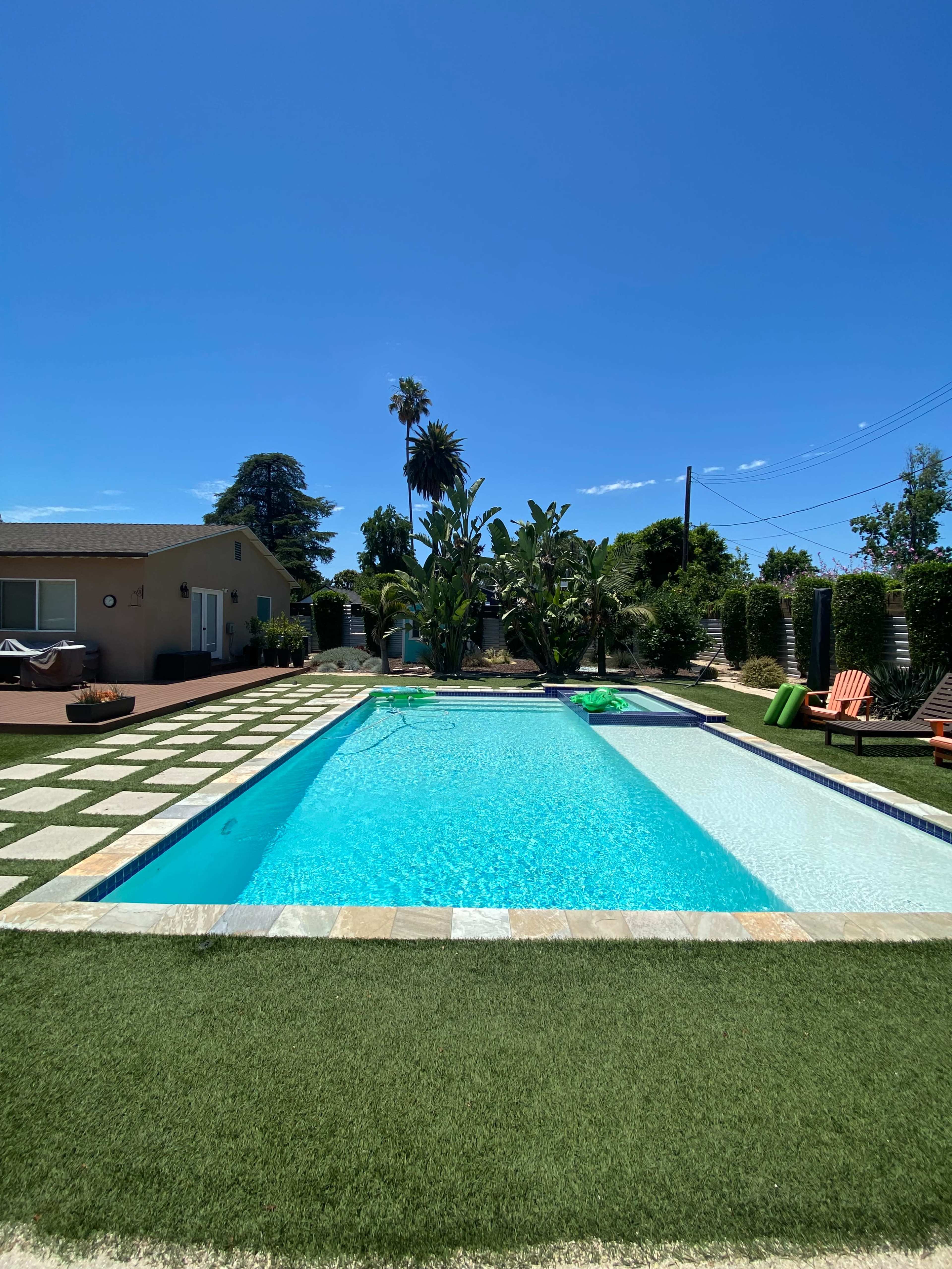 The image shows a clear swimming pool surrounded by lush greenery and a checkered stone patio under a bright blue sky.