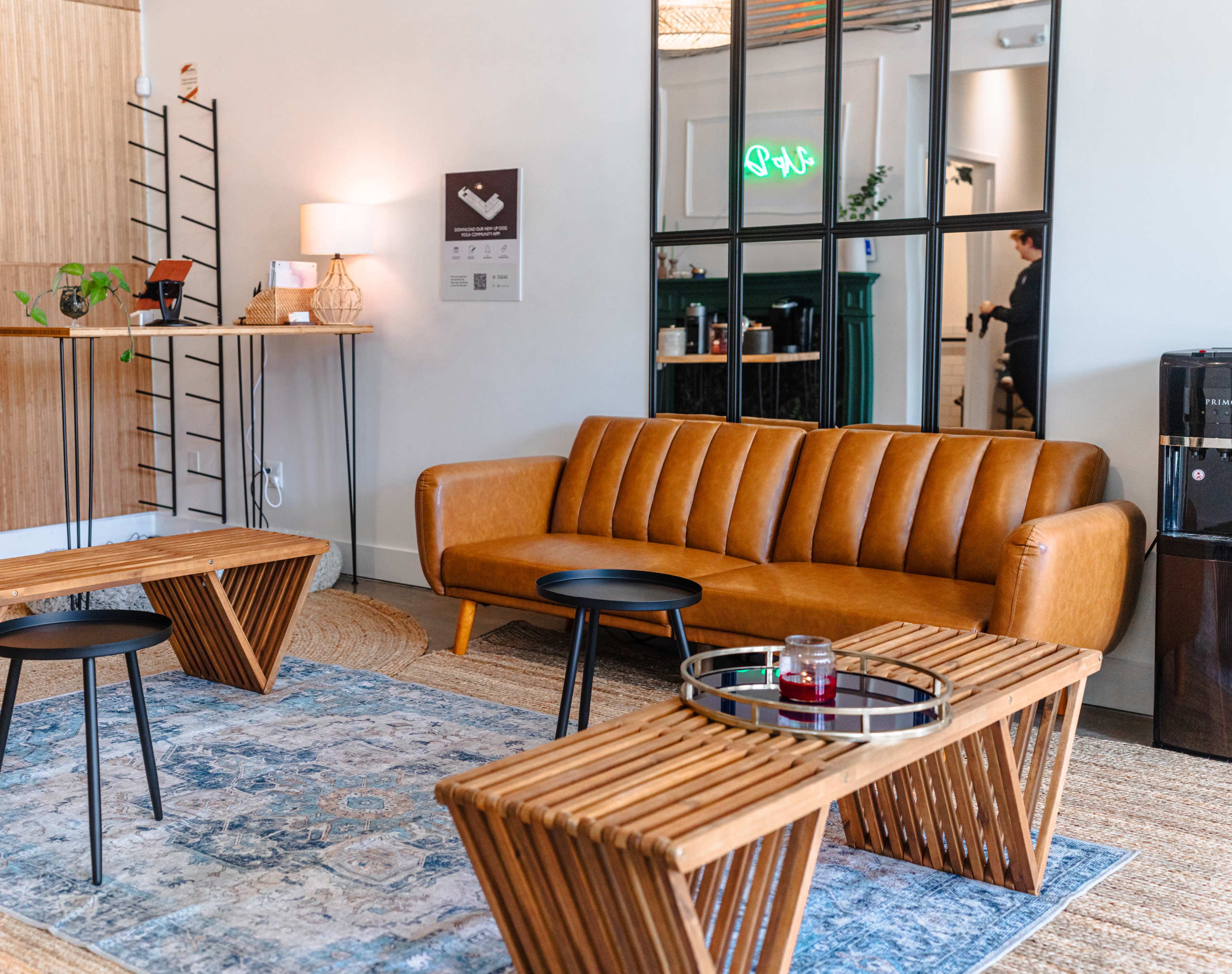 A modern lounge area features a tan leather couch, wooden coffee tables, and a decorative rug, with a glass partition reflecting a hallway.