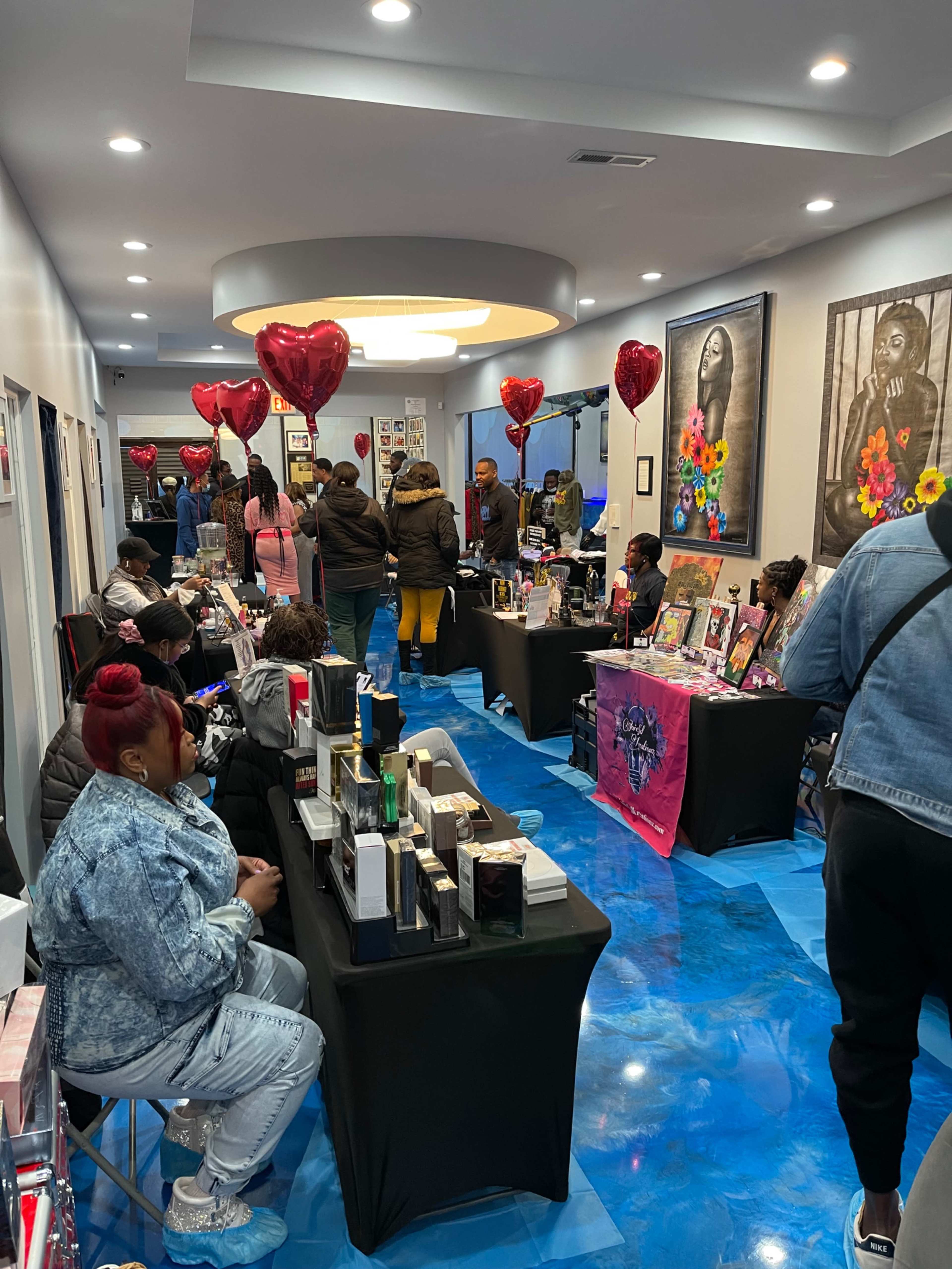 A vibrant indoor market features multiple booths with vendors selling various products while attendees browse, surrounded by heart-shaped balloons.