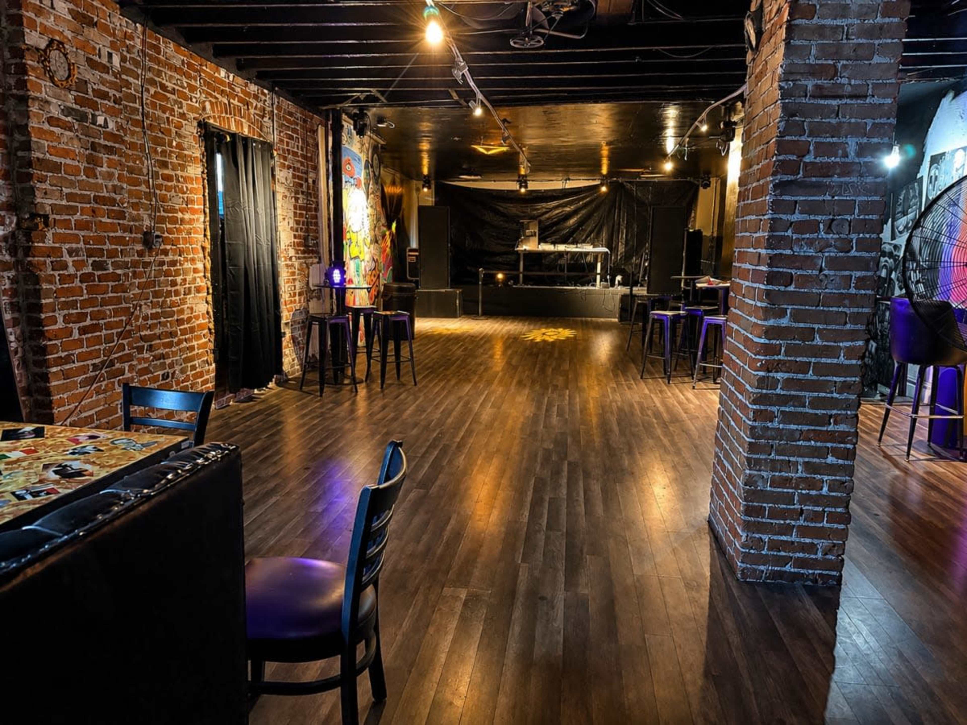 The image shows an empty venue with brick walls, a dark stage area, and wooden flooring, featuring scattered tables and chairs.