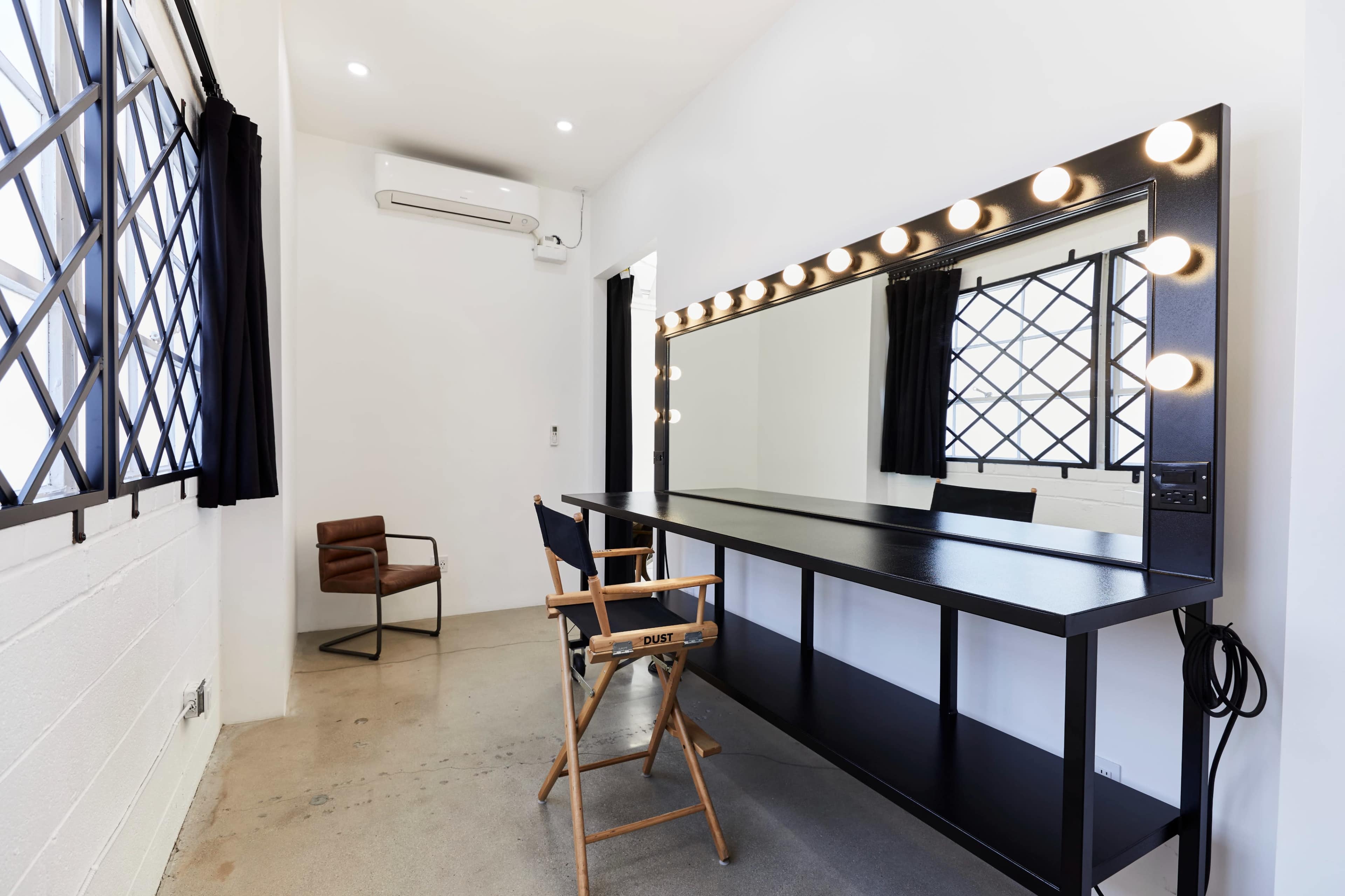 A minimalist makeup room featuring a large mirrored vanity with lights, a director's chair, and a contemporary seating area by the window.