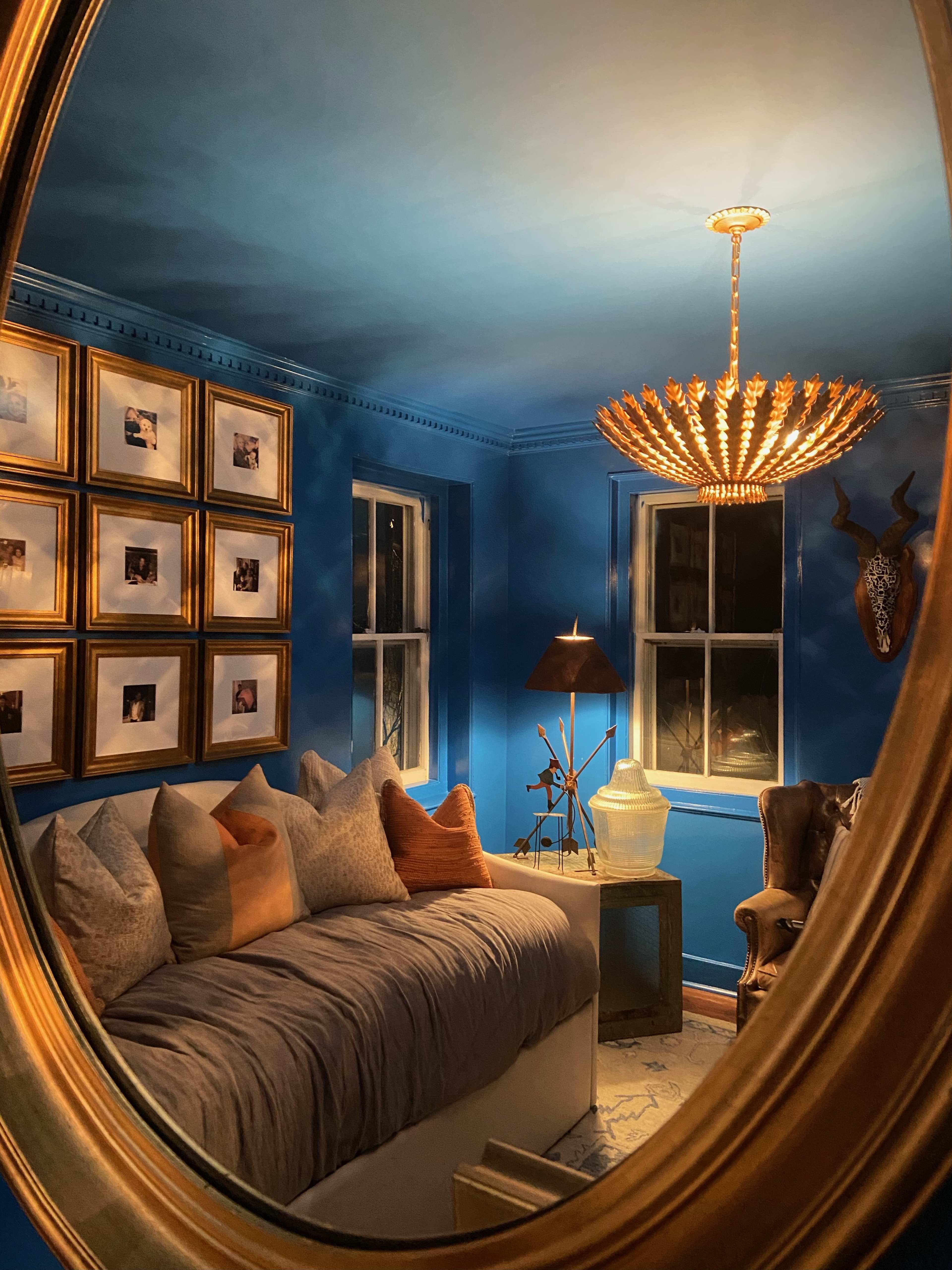 The image reflects a cozy bedroom with blue walls, a gallery of framed photos, and a modern chandelier above a daybed.