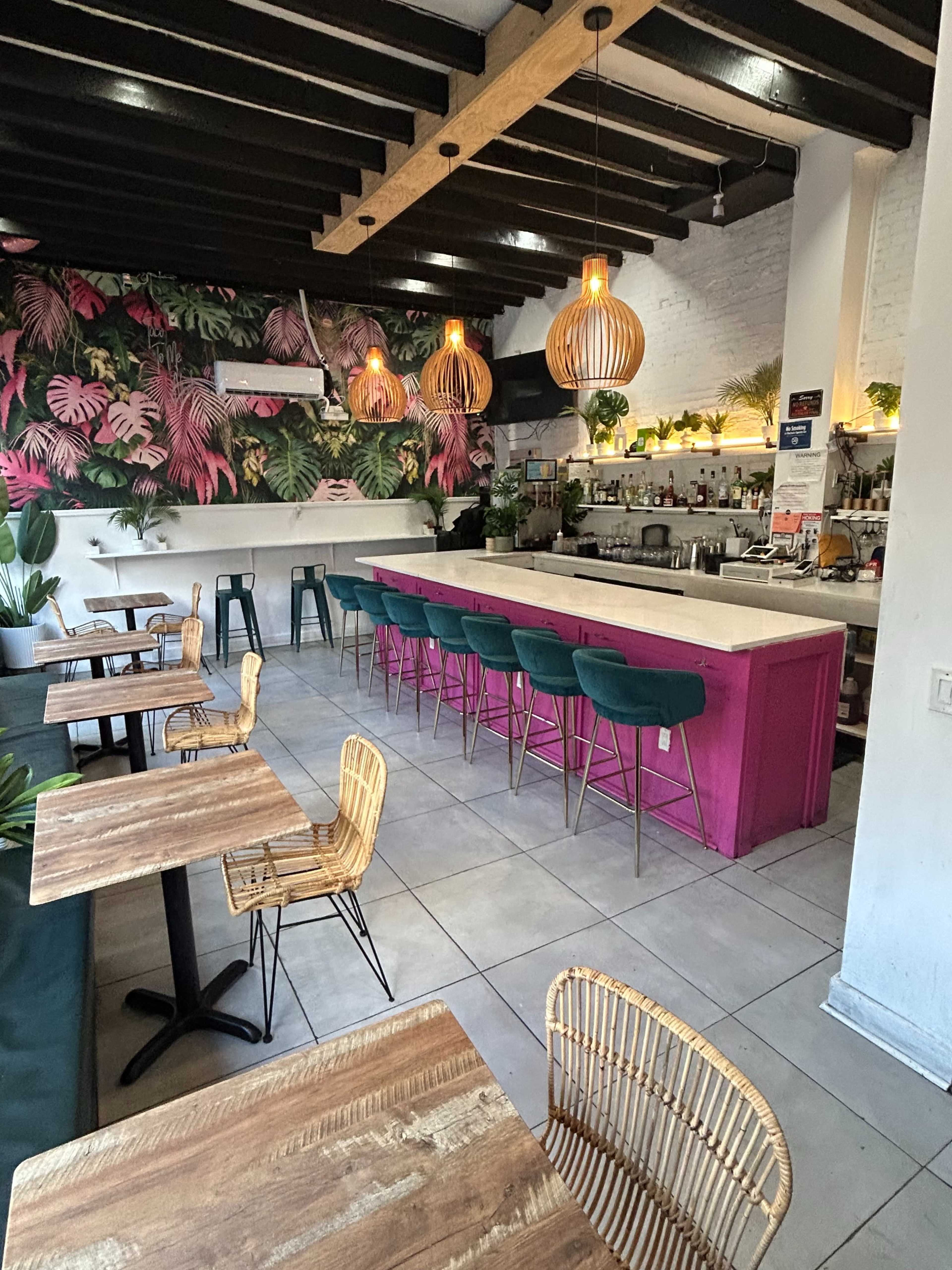 The image shows a modern cafe with a colorful tropical wallpaper, wooden tables and chairs, and a pink bar area illuminated by pendant lights.