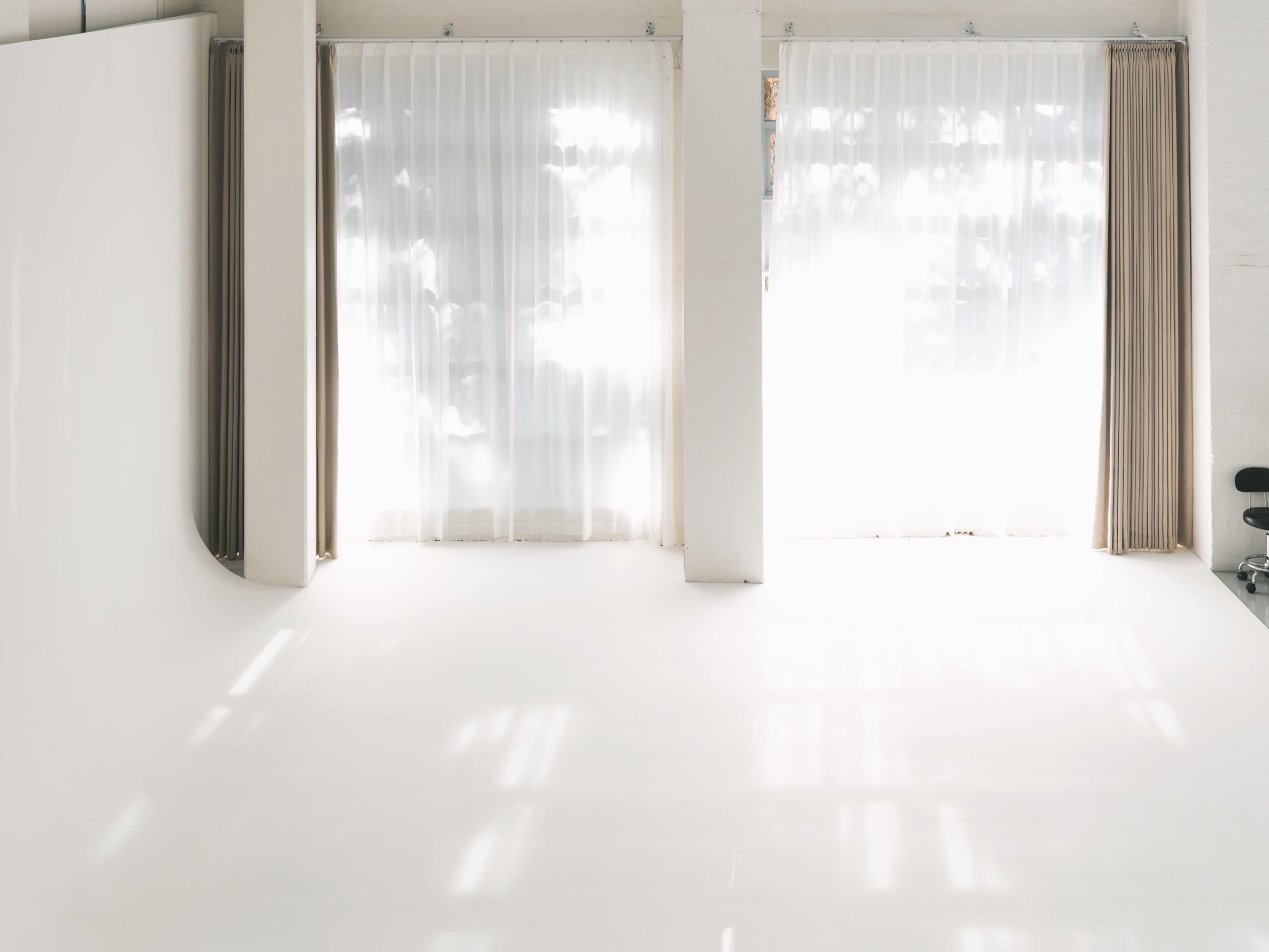 The image shows a bright, empty studio space with white walls and sheer curtains allowing natural light to enter.