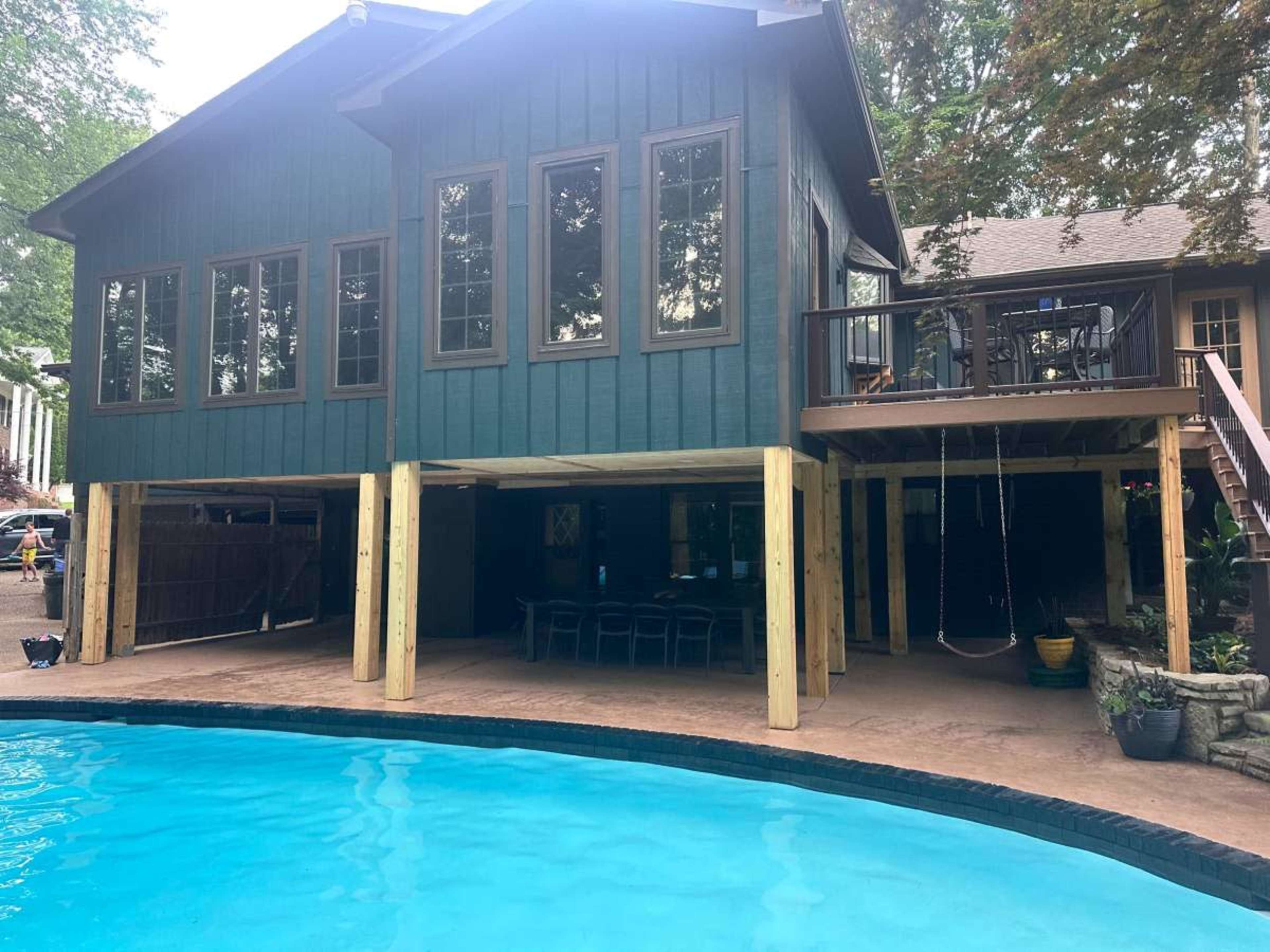 A two-story house with a wooden exterior is elevated above a pool, featuring large windows and a covered outdoor seating area underneath.