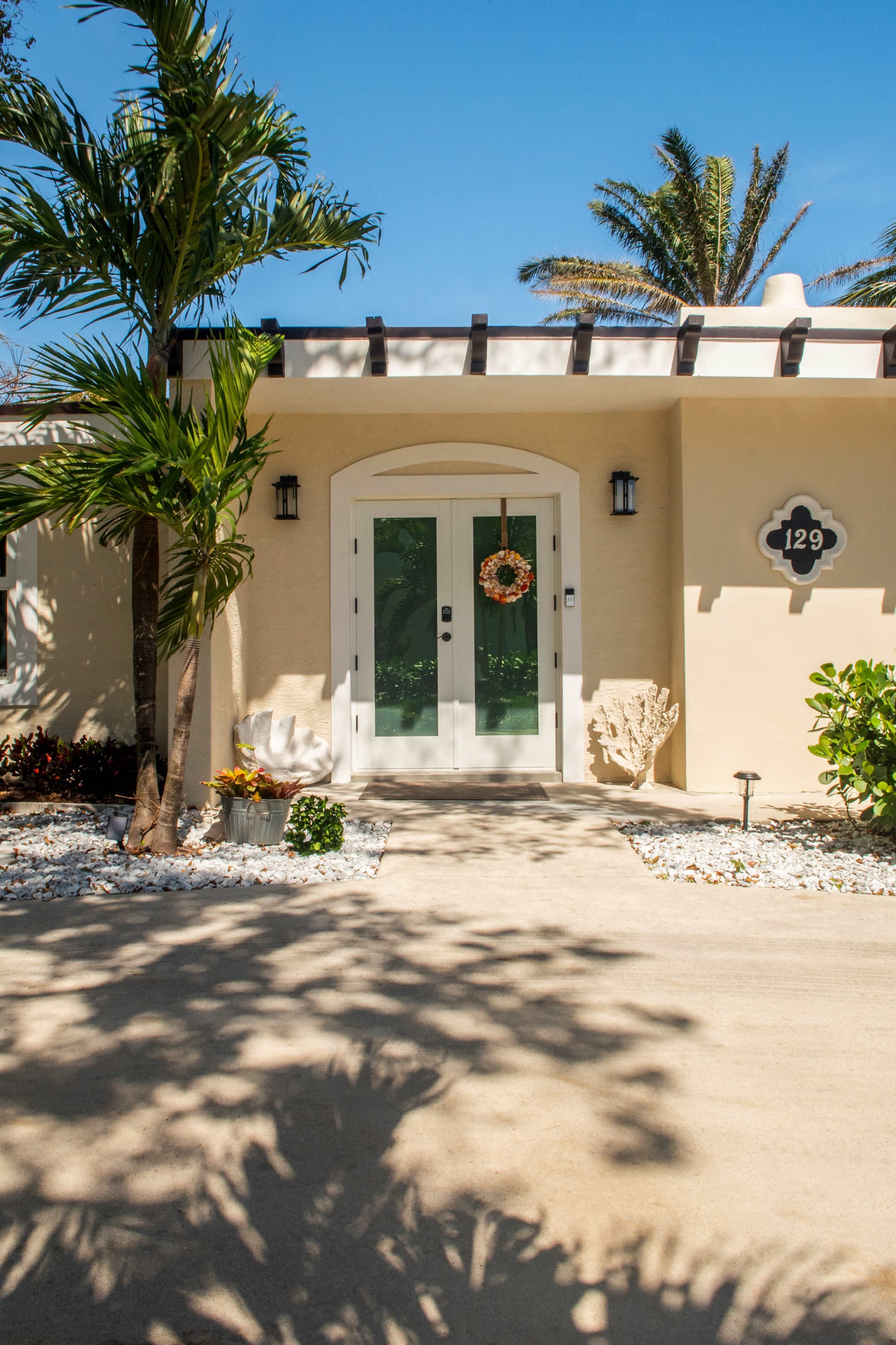The image shows a light-colored home entrance with double doors adorned by a wreath, flanked by palm trees and landscaped greenery.