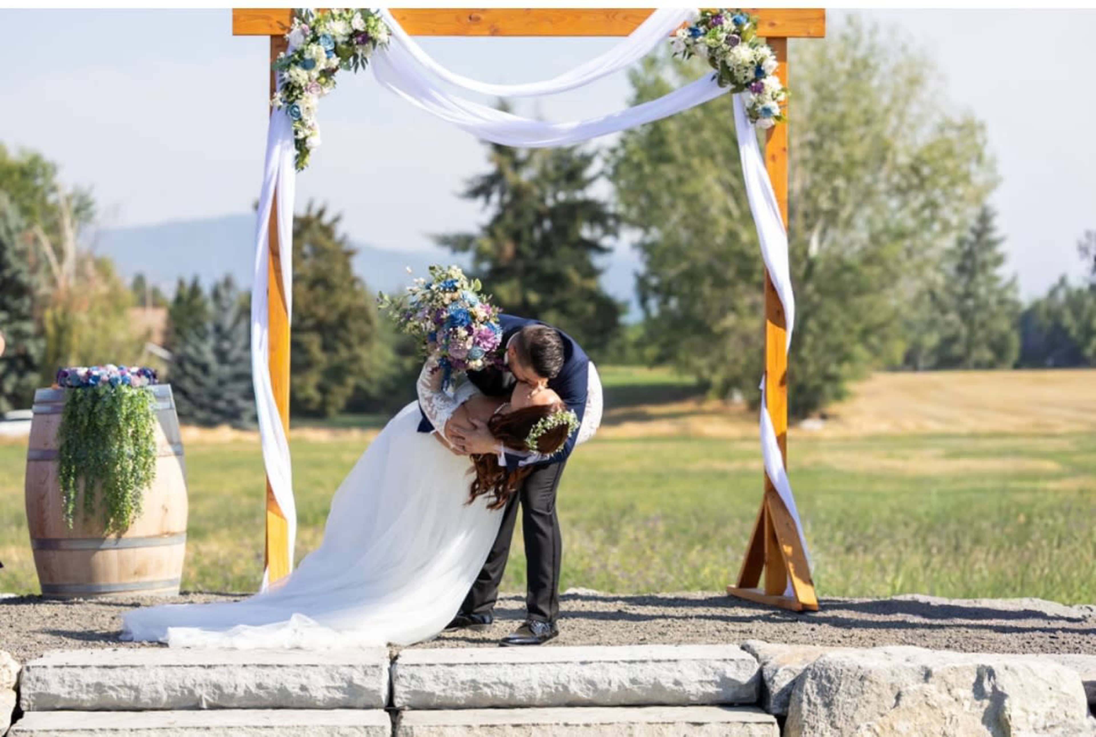 A couple embraces and shares a kiss under a decorated wedding arch in a scenic outdoor setting.