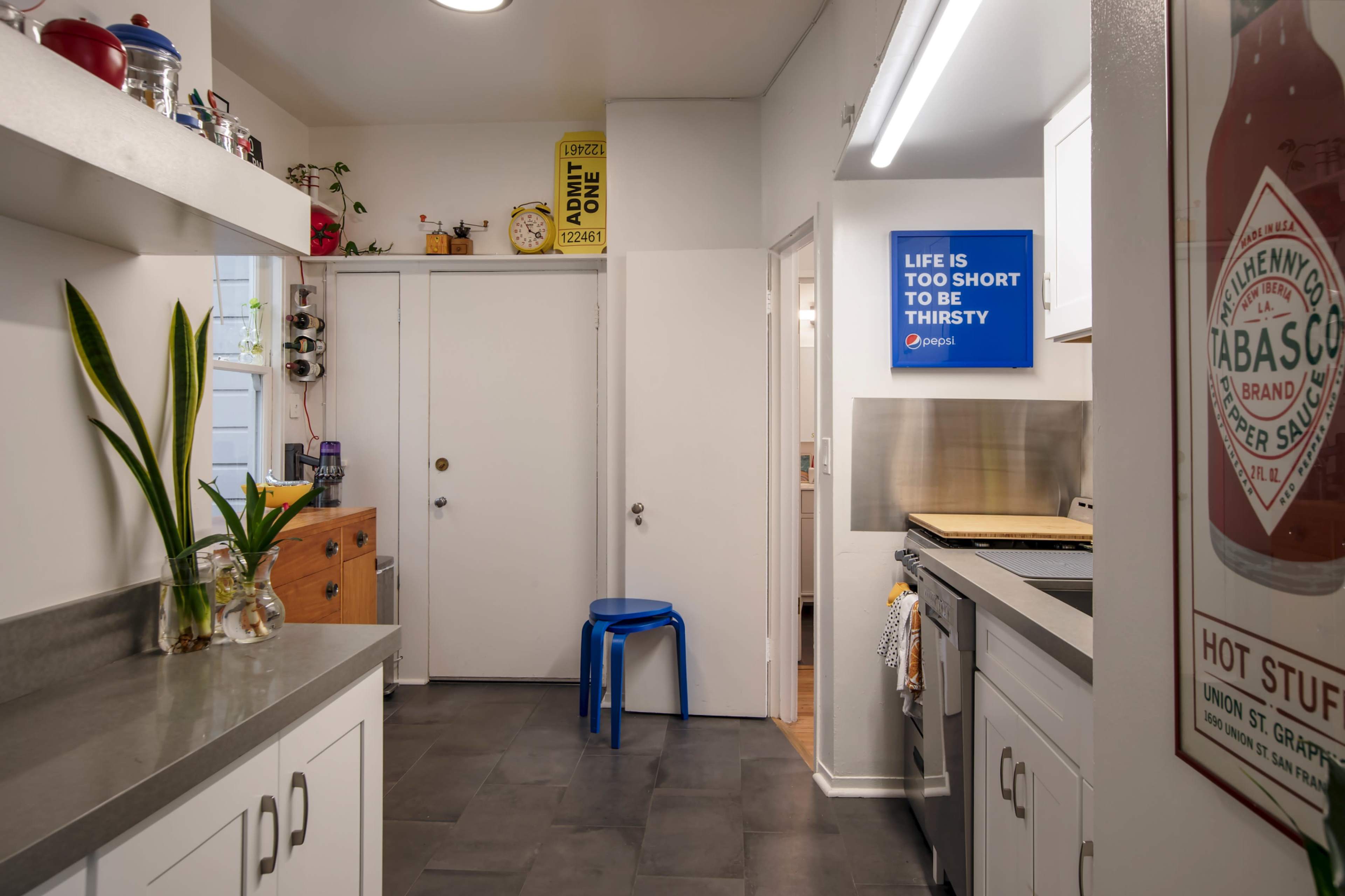 A kitchen with grey countertops, a blue stool, a door leading to another room, and a wall-mounted sign that reads "LIFE IS TOO SHORT TO BE THIRSTY."