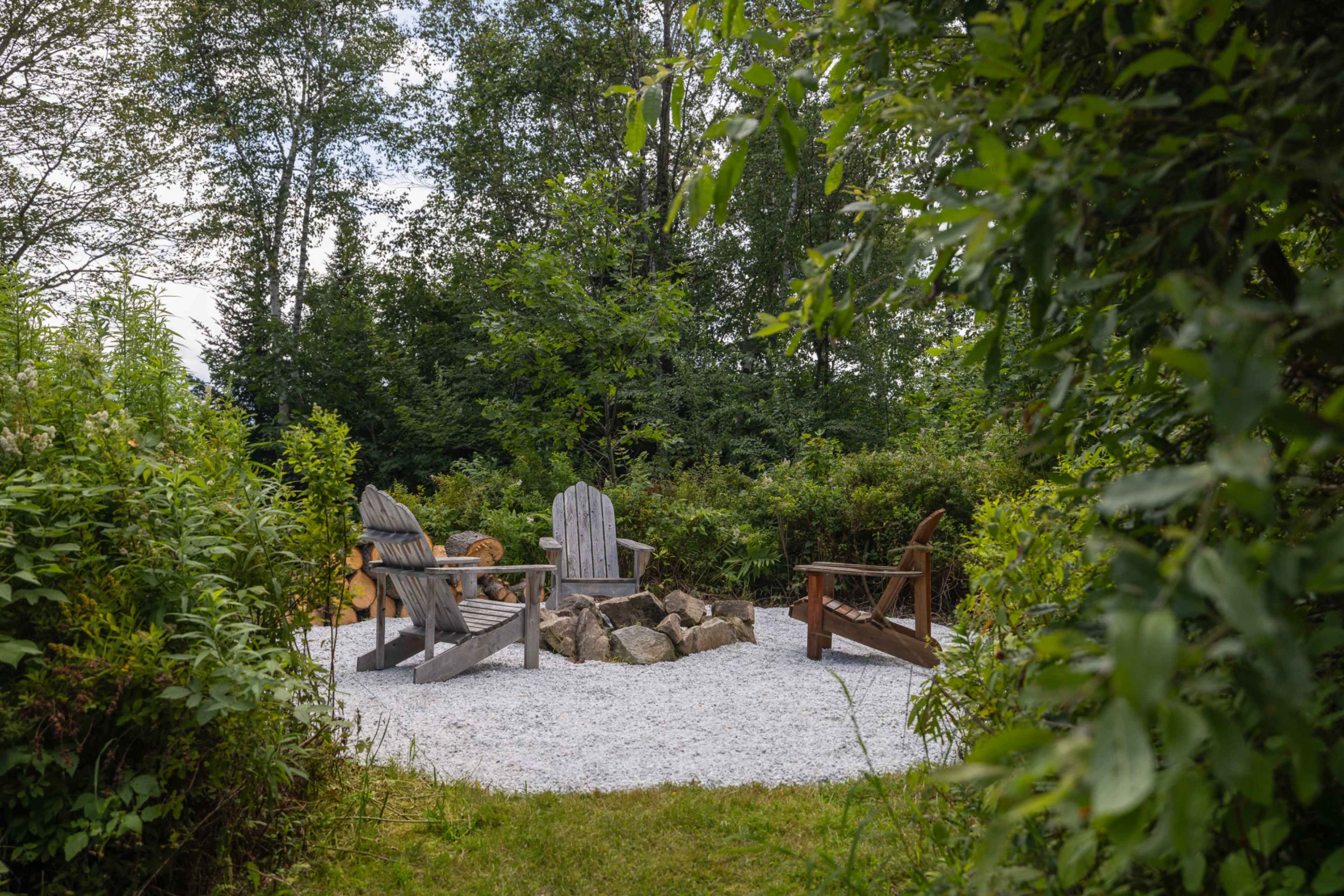 A small gravel seating area with four wooden chairs is surrounded by lush greenery and trees.