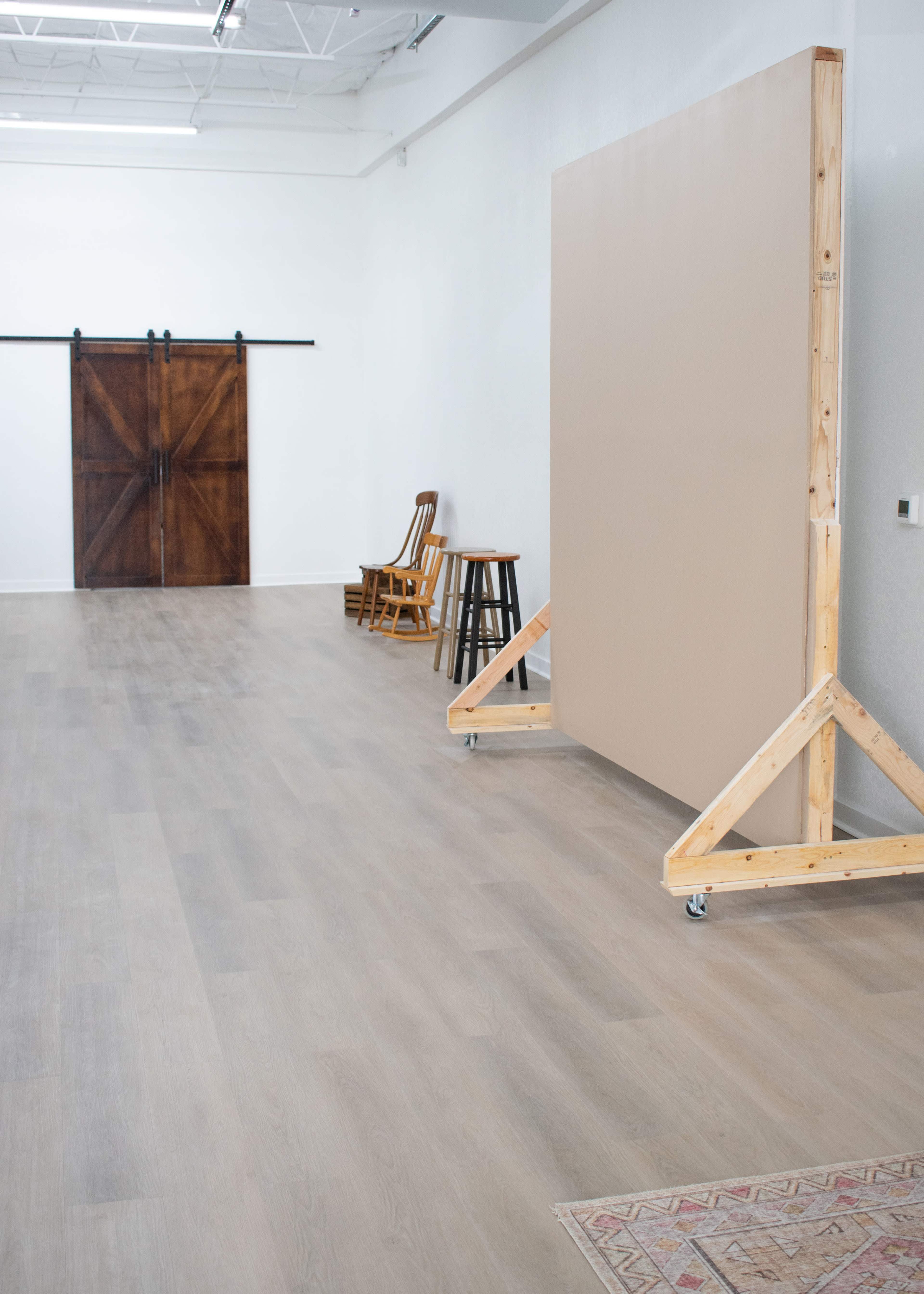 A spacious interior with wooden flooring, featuring a large, movable beige panel, three wooden chairs, and a set of sliding barn doors in the background.