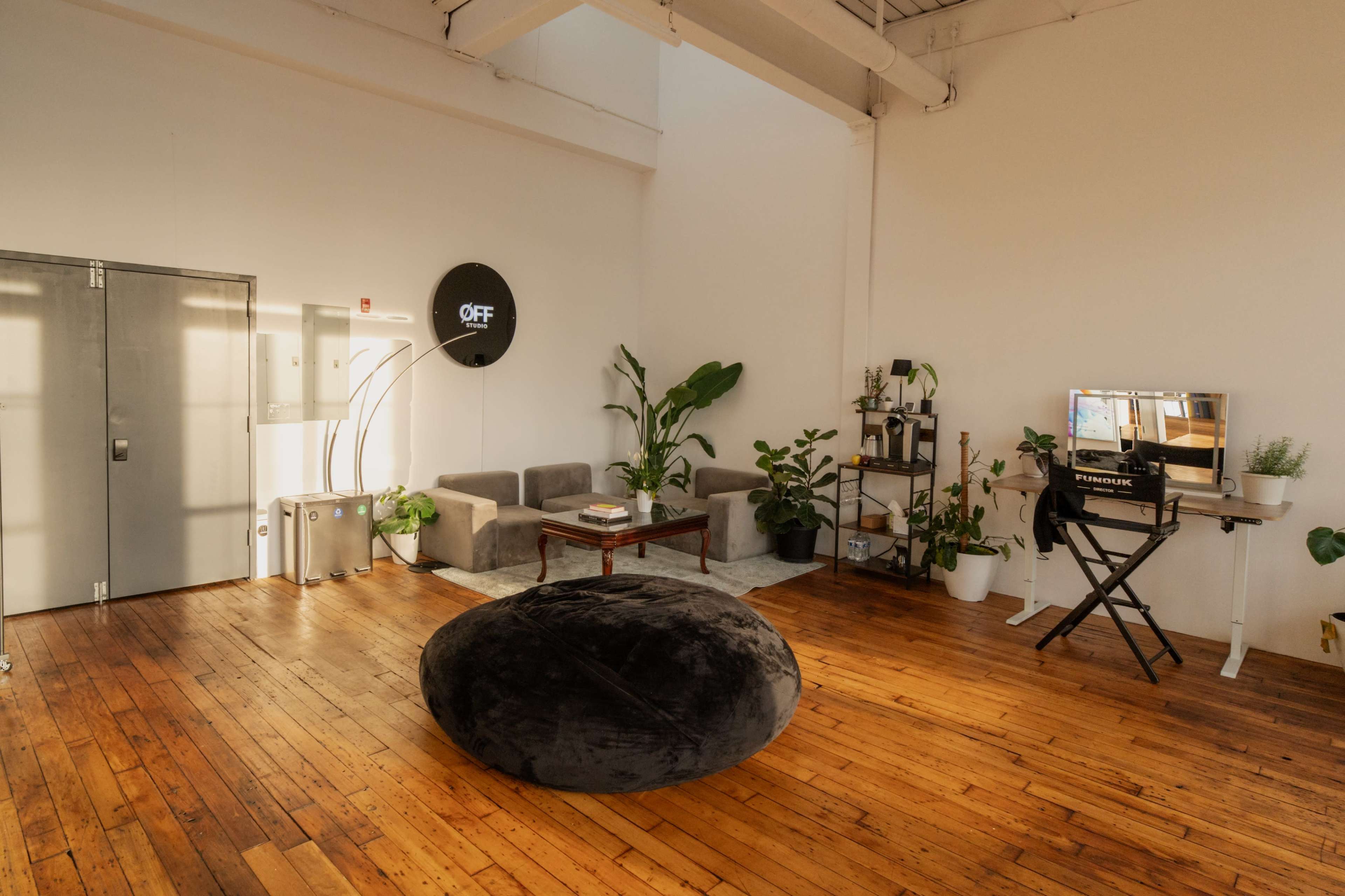 A minimalist interior features a large black bean bag, a seating area with gray sofas, a small table, potted plants, and a desk with a monitor against white walls and wooden flooring.